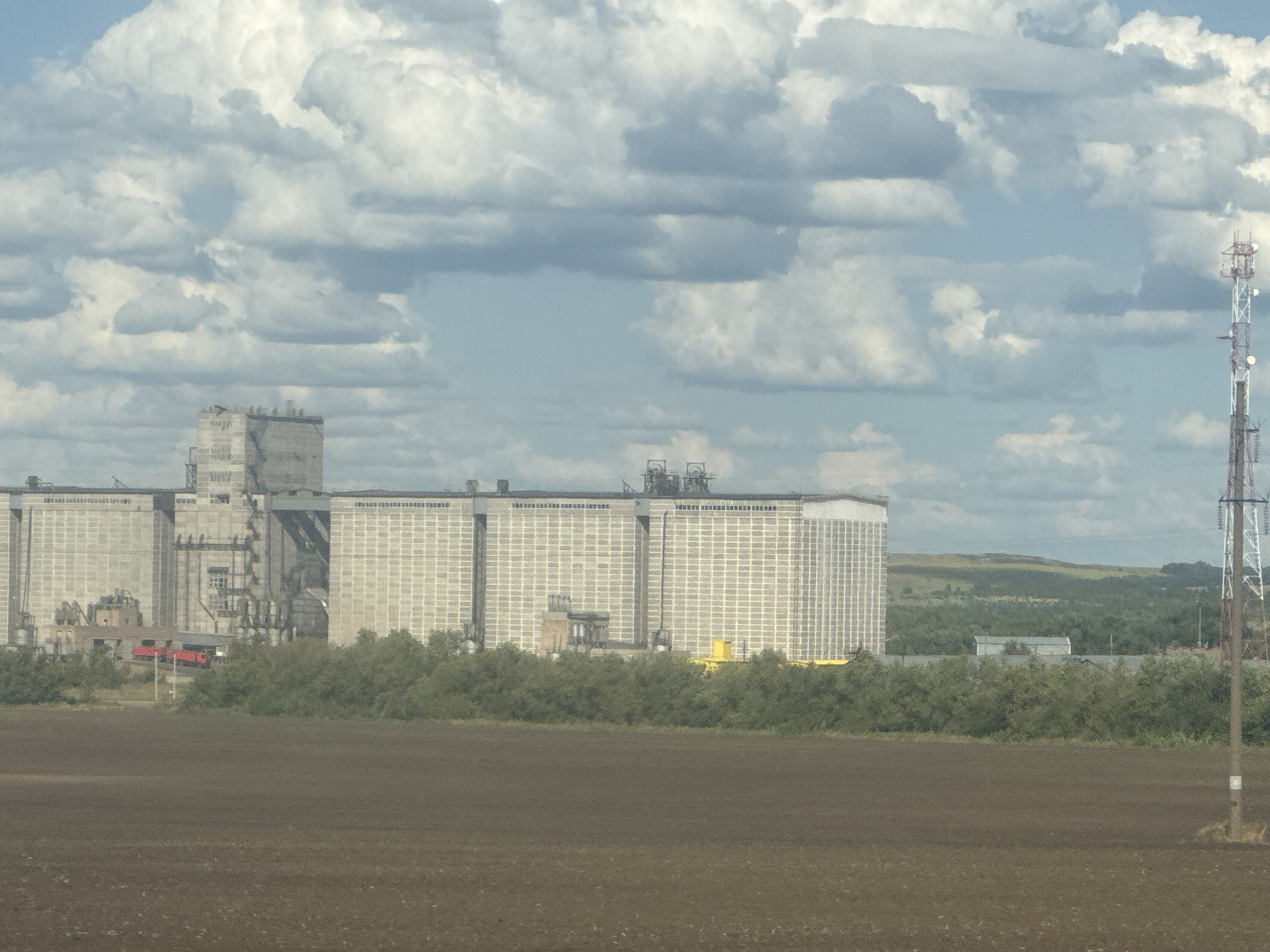This is a distant, wide shot of a large industrial facility, likely grain elevators, in Novosergiyevka, Russia. The facility is characterized by a row of tall, rectangular buildings with a grid-like facade, constructed of concrete or similar material. To the left, a section of the structure appears more complex, with visible pipes, platforms, and scaffolding. A small, red train car is visible at the base of the facility, possibly loading or unloading. The buildings are set against a backdrop of green trees and rolling hills. In the foreground, a vast, dark brown, tilled field stretches across the lower half of the image. A tall, lattice-like communication tower stands to the right of the industrial complex. The sky is filled with large, puffy white and gray clouds, suggesting a partly cloudy day. No people are discernible in the image.