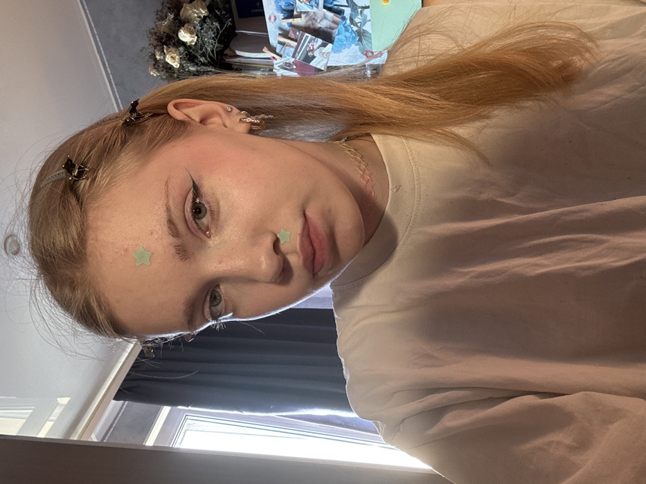 The image presents a young woman with light skin and blonde hair, partially secured by two dark hair clips on the right side of her head, with the remainder flowing over her right shoulder. Her green eyes are directed towards the viewer. She exhibits winged eyeliner and pink blush on her cheeks. Two light green, star-shaped stickers are applied to her face: one centered on the forehead between the eyebrows, and a second on the philtrum above her upper lip. Her right ear features multiple piercings, specifically a small stud in the upper cartilage and two hoops in the lower lobe, one of which includes decorative elements. She wears a translucent, multi-colored choker necklace and a plain white, loose-fitting crew-neck t-shirt.
The scene takes place indoors, within what appears to be a residential room. To the left, a window provides natural illumination, with dark curtains partially drawn. To the right, in the upper portion, a dried floral arrangement consisting of light-colored flowers and foliage is visible. Below this, several photographs or postcards are attached to a light-colored vertical surface with wooden clothespins; some of these items display blue and white patterns. A segment of a light green object is discernible in the lower right corner of the frame.
