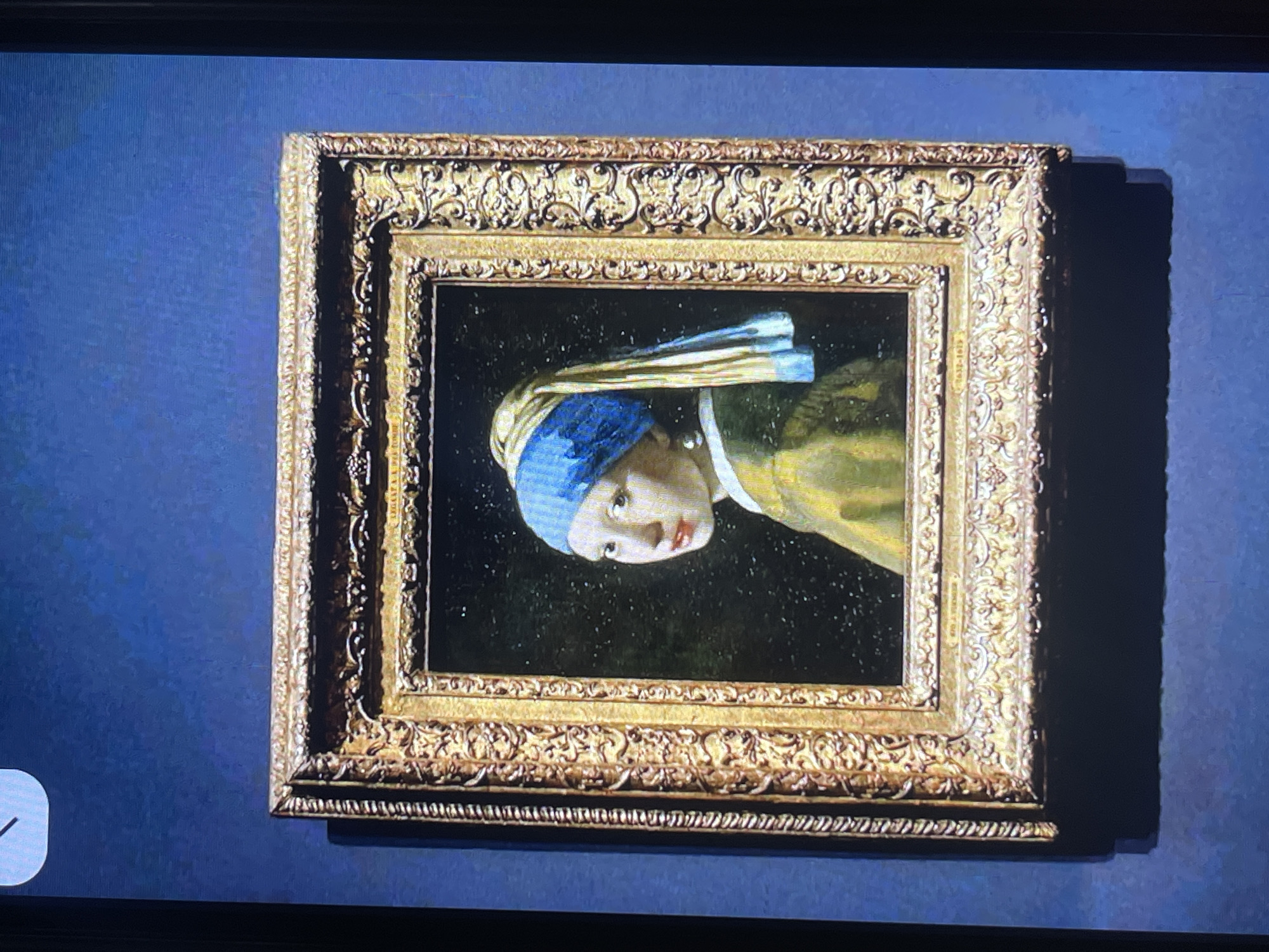The image displays Johannes Vermeer's famous painting, "Girl with a Pearl Earring," displayed in a richly ornate golden frame. The painting itself is centered within the frame and depicts a young woman from the shoulders up, gazing to her left. She wears a blue and yellow headscarf and a simple brown garment, with a prominent pearl earring adorning her left ear. The background of the painting is dark and nondescript, making the subject stand out. The frame exhibits intricate scrollwork and floral motifs characteristic of Baroque art.
The painting is hung on a dark blue wall, which appears to be the background of a museum or gallery setting. To the upper left of the framed painting, a portion of a white clock face with black hands is visible, suggesting the presence of a timekeeping device within the same space. The overall lighting suggests an indoor environment, likely a well-lit exhibition hall. There is a distinct shadow cast by the bottom of the framed painting onto the wall, indicating a light source from above or the front.
The specific location in Bishkek City, Kyrgyzstan, is not discernible from the image itself, as it lacks any identifiable landmarks or contextual clues of that city. The image captures only the artwork and its immediate display environment.