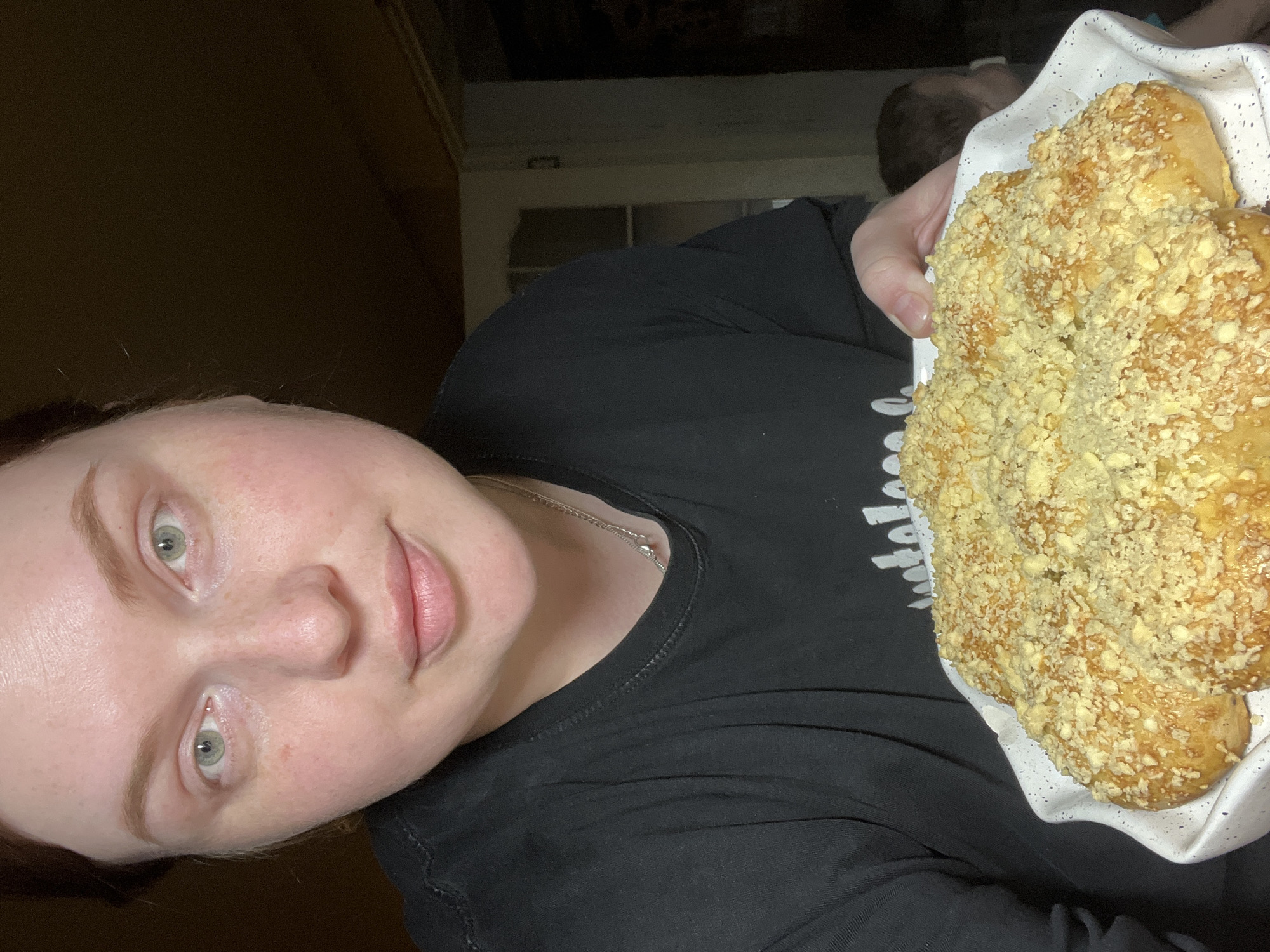 A person with pale skin and green eyes is holding a plate of sweet bread in a dimly lit indoor location, possibly in a home. The bread is covered in a light-colored crumble topping. The person is wearing a black t-shirt and a silver necklace. A doorway is visible in the background. There is no information to confirm the location is Павловка, Russia.