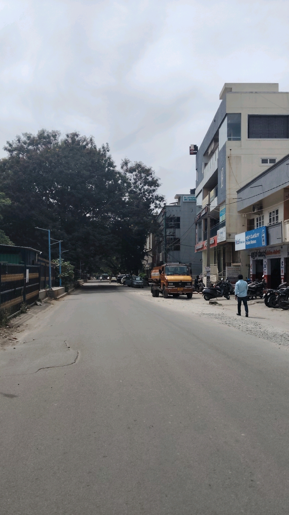 The image depicts a street scene in Bengaluru, India. The road is paved and appears to be in moderate condition, with some cracks visible. On the left side of the street, there is a tall, dense tree providing shade and a black metal fence with yellow accents. Further down the street, several cars are parked.
On the right side of the street, there are multi-story buildings with commercial establishments on the ground floors. A yellow truck is parked in the middle of the road. Several motorcycles are parked along the sidewalk in front of the buildings. A man in a light blue shirt and dark pants is standing on the sidewalk, facing away from the camera. The sky is overcast. The overall impression is that of a typical urban street on a cloudy day in Bengaluru.