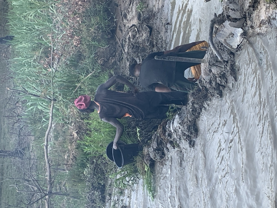 Two individuals stand by a muddy riverbank. One, wearing a maroon head covering, black shirt, and orange undershirt, bends while holding a black bucket. The other person stands with a tool and is clad in a black shirt with a yellow undershirt. Mud, grass, and various plants surround the riverbank. Water flows in the river.