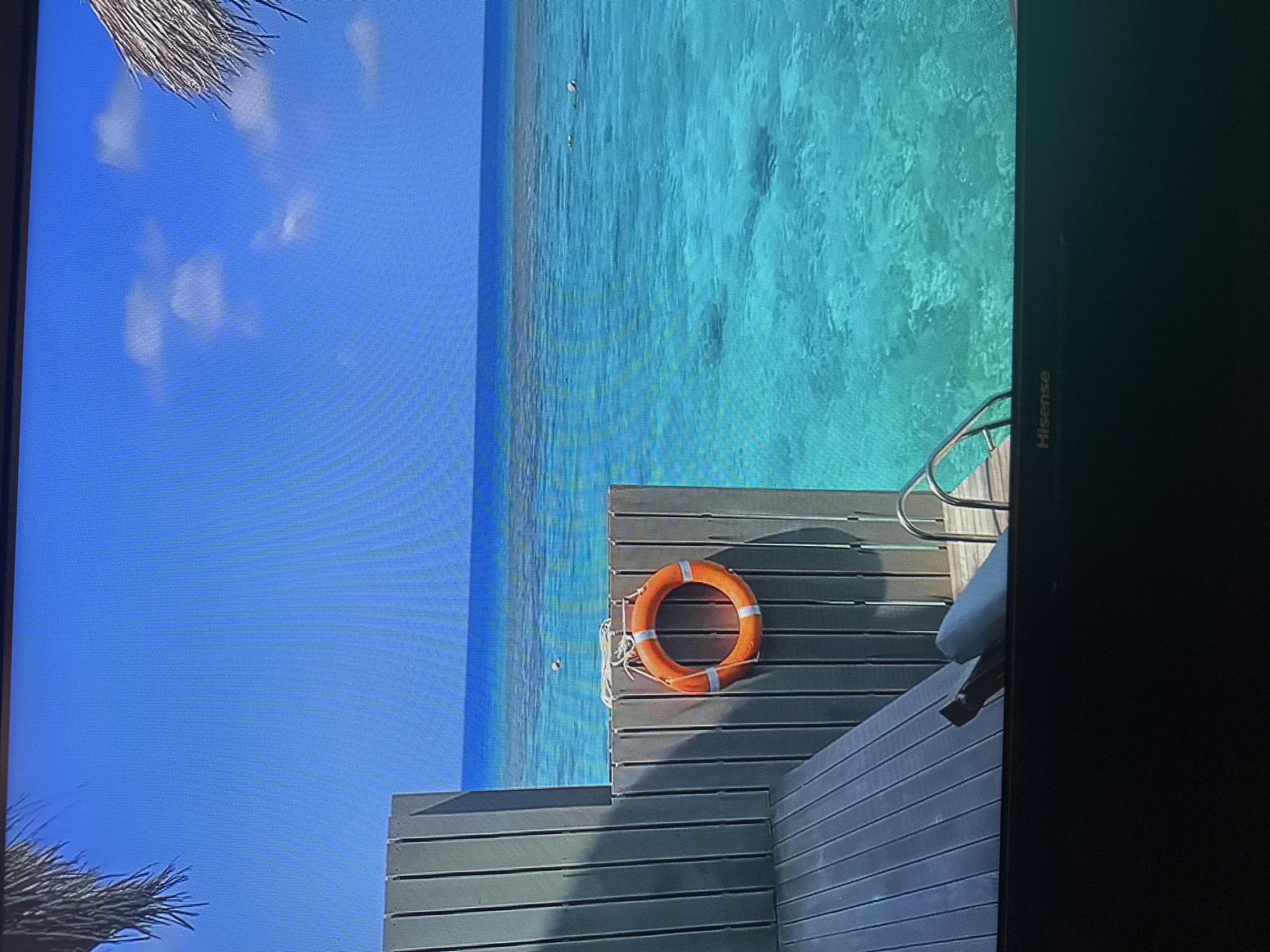This is an image of a tropical scene, likely a resort, with clear turquoise water and a blue sky. There is a wooden deck with a life preserver hanging on a vertical wooden fence. A metal ladder leads down into the water. In the distance, there are coral reefs visible beneath the surface of the water and a few people can be seen swimming or snorkeling. There is also a thatched roof structure visible at the top right of the image. The scene is peaceful and relaxing. There is no indication of the city of Kyiv, Ukraine, or any urban elements in this image.