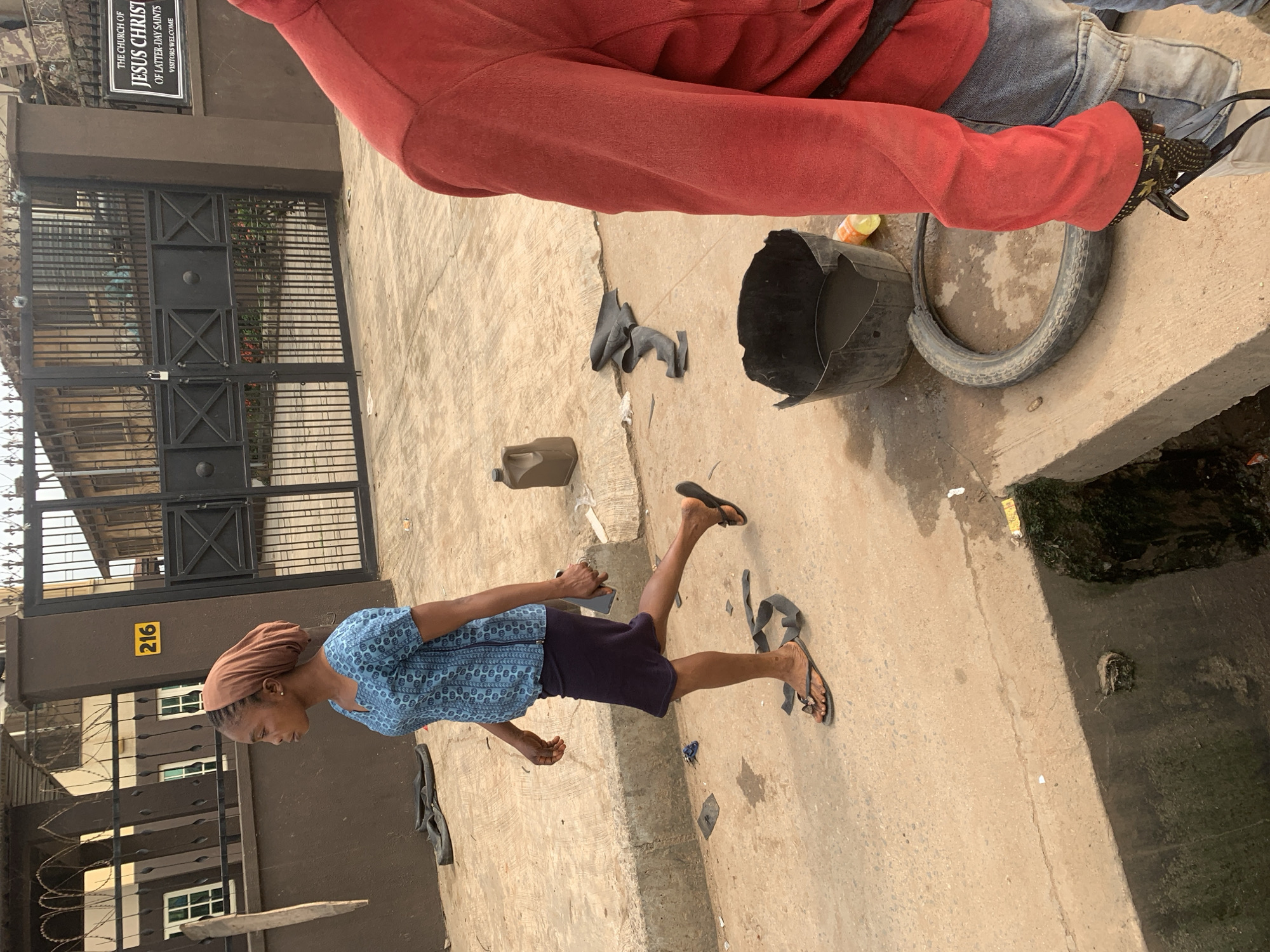 A medium shot captures a young woman walking from left to right across a street in Shagamu, Nigeria. She has dark skin and wears a light brown headscarf wrapped around her hair. Her blue short-sleeved shirt features a repeating pattern of light blue geometric shapes. She is also wearing a dark purple knee-length skirt. The woman's feet are clad in dark flip-flops. Her left arm is bent at the elbow, and her right hand is holding a mobile phone.
The background reveals a brown building with black metal gates and a sign that reads "THE CHURCH OF JESUS CHRIST OF LATTER-DAY SAINTS VISITORS WELCOME." A yellow number "216" is affixed to the building. The street pavement is concrete. Scattered on the ground are various objects, including a dark plastic container, a broken black bucket with water, a deflated tire, and other discarded items. To the right of the frame, the arm and shoulder of another person are visible, wearing a red long-sleeved shirt and blue jeans.