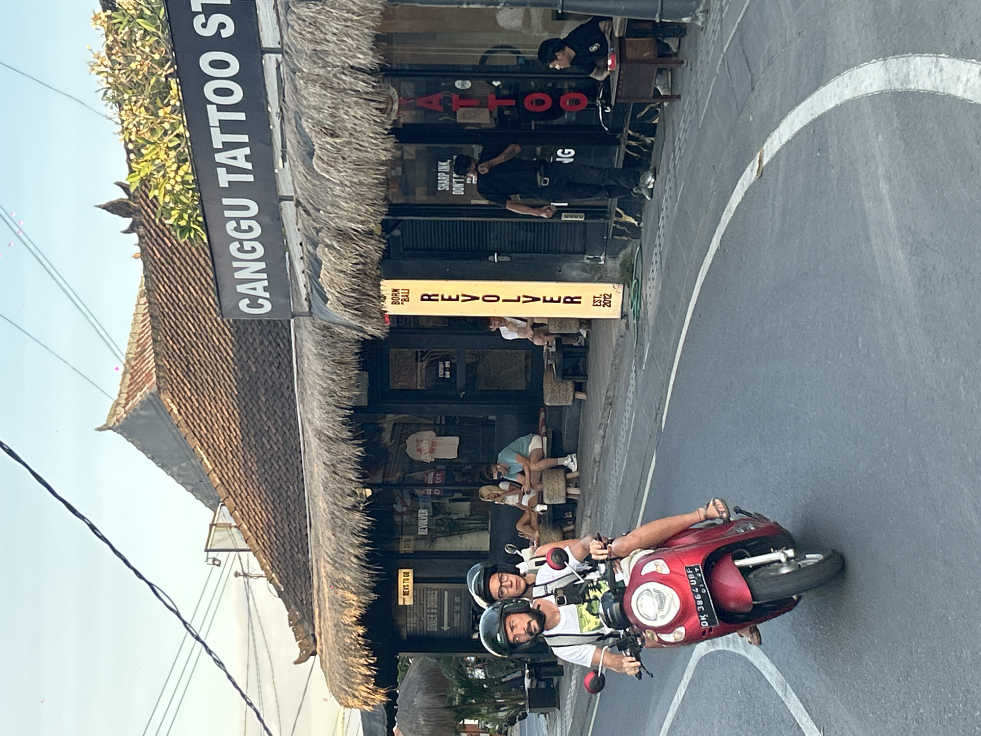 A man and a woman wearing helmets are riding a red scooter on a paved road in North Kuta, Indonesia. The man is driving, and the woman is sitting behind him. The scooter has a license plate that reads "DK 3864 UBF". In the background, there is a building with a thatched roof that houses a tattoo parlor called "Canggu Tattoo Studio". Several people are visible outside the tattoo parlor, some sitting on stools and others standing. The environment appears to be a street scene in a tropical location, with palm trees and other vegetation visible in the distance. The sky is clear and blue.