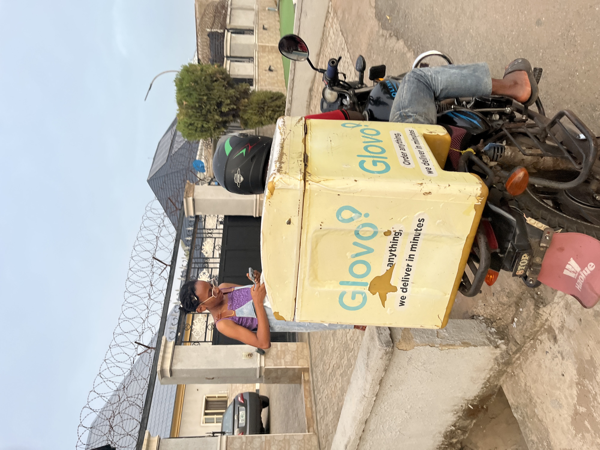A Glovo delivery person sits on a motorbike on a paved street in front of a gated property in Abuja, Nigeria. The delivery person, a young woman, is wearing light-wash denim overalls over a purple top and gold-rimmed glasses. She is looking down at her phone in her hand. The delivery box attached to the back of the motorbike is yellow and branded with the Glovo logo and tagline "Order anything, we deliver in minutes". A black helmet sits on top of the box. The motorbike is black and the seat appears to have faded blue fabric. The building behind her has a dark grey metal gate with razor wire. A black car is visible behind the gate. Other buildings and greenery can be seen in the background.