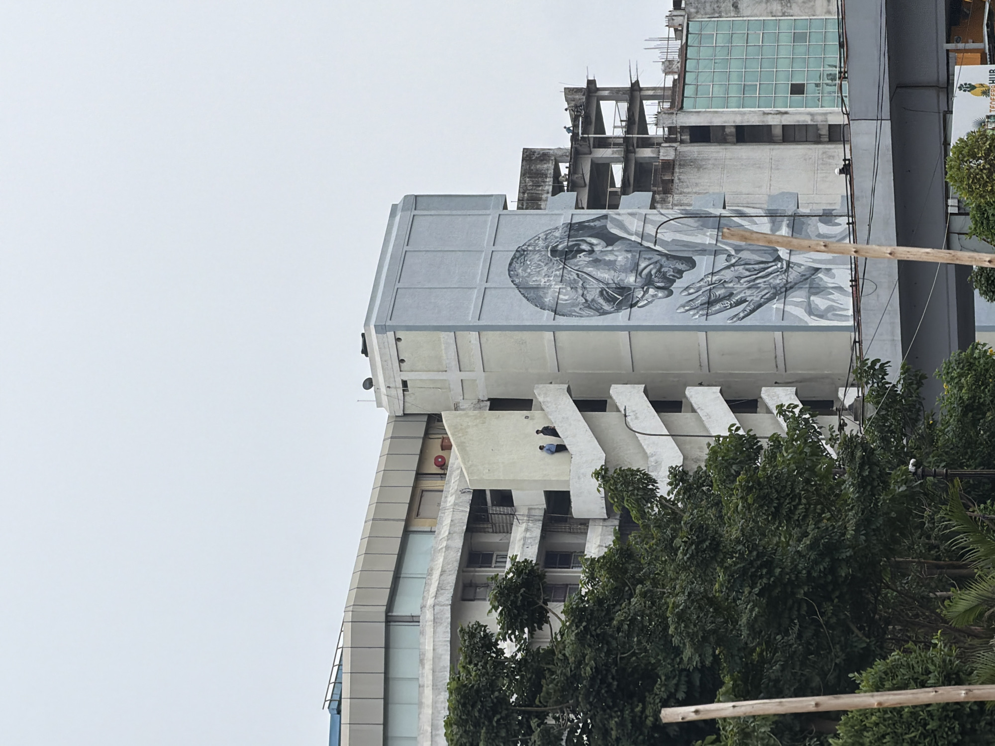 The image depicts a large mural of Mahatma Gandhi praying, painted on the side of a building in Hyderabad, India. The mural is a monochrome portrait, focusing on Gandhi's face and hands. To the left of the mural, a multi-story building with balconies and an external staircase is visible, with two individuals standing on the staircase. Further to the right, an unfinished concrete structure is under construction, and a section of a building with blue-tinted windows is also present. In the foreground, lush green trees partially obscure the lower part of the buildings. The sky is overcast, suggesting a cloudy day. The overall scene portrays a blend of urban development, public art, and ongoing construction.