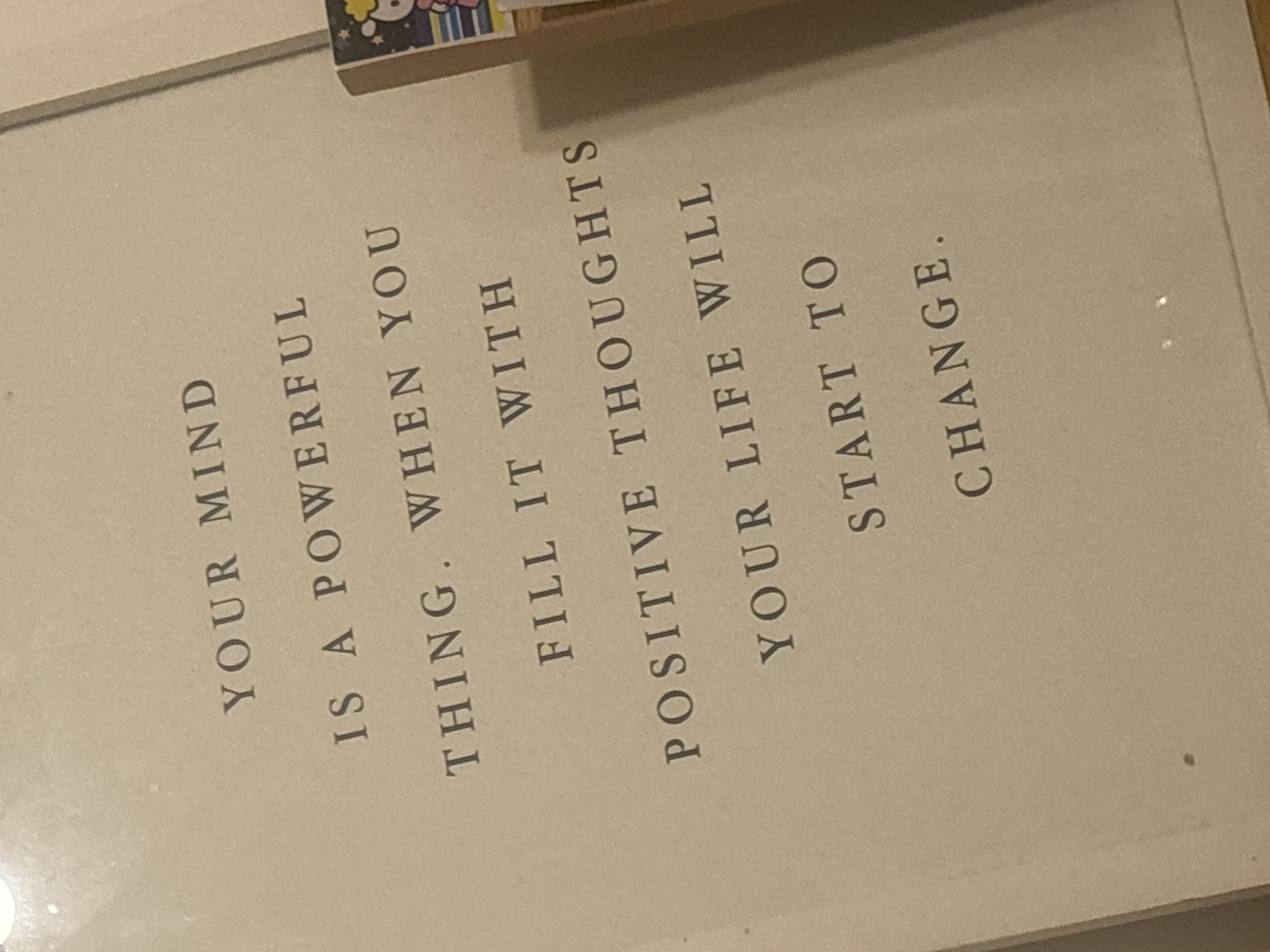 An indoor photograph displays a framed motivational print, set at an angle. The print features a quote in a dark serif font on a light, off-white background. Partially visible on the top edge of the frame is a small, colorful decorative object, possibly a clip or magnet, depicting cartoon-like characters.
The text on the print reads:
"YOUR MIND
IS A POWERFUL
THING. WHEN YOU
FILL IT WITH
POSITIVE THOUGHTS
YOUR LIFE WILL
START TO
CHANGE."
The lighting suggests an indoor setting, possibly a room or office. There are no people visible, and no specific activity is taking place; the image captures a static display. While no distinct visual cues for the location are present within the frame, the scene is situated in Buenos Aires, Argentina. The overall message of the print is one of empowerment and positive thinking.