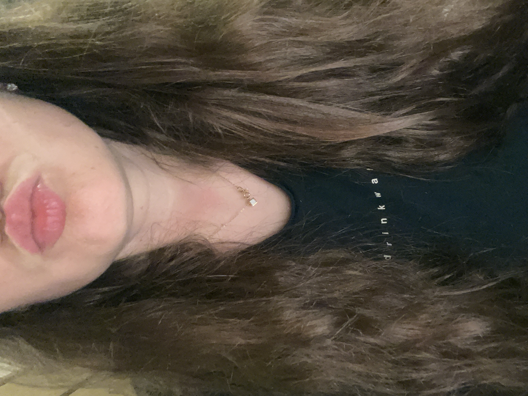 A close-up, chest-up, medium shot shows a woman with long, wavy, brown hair framing her face. She is wearing a dark green t-shirt with white text that reads "drink wa". A delicate rose gold necklace with two small, interconnected rings and a diamond accent is visible around her neck. Her lips are parted in a pout, and she is wearing subtle makeup. A small, sparkling stud earring is visible in her right ear. The background is indistinct and appears to be an indoor setting.