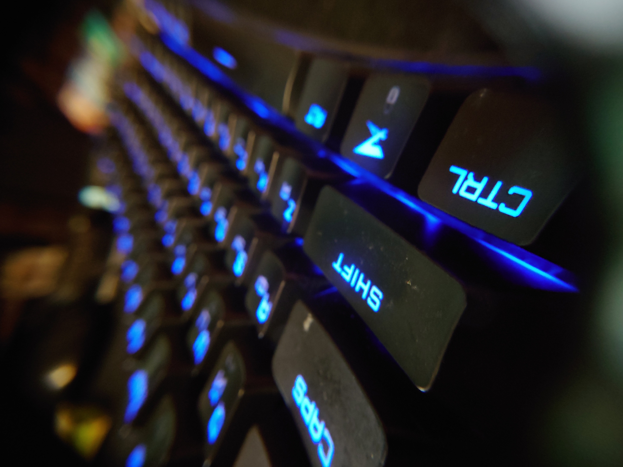 This is a close-up, low-angle shot of a black mechanical keyboard with blue backlighting. The focus is on the keys, with some appearing in sharp focus while others are blurred due to the shallow depth of field. Visible keys include "CAPS", "SHIFT", and "CTRL", all illuminated by the blue LEDs. The overall scene is dark, with the blue light from the keyboard being the primary light source. The background is out of focus, showing blurred shapes and colors that suggest a dimly lit room. There is no indication of the city of Sokolohirsk, Ukraine, in the image.
