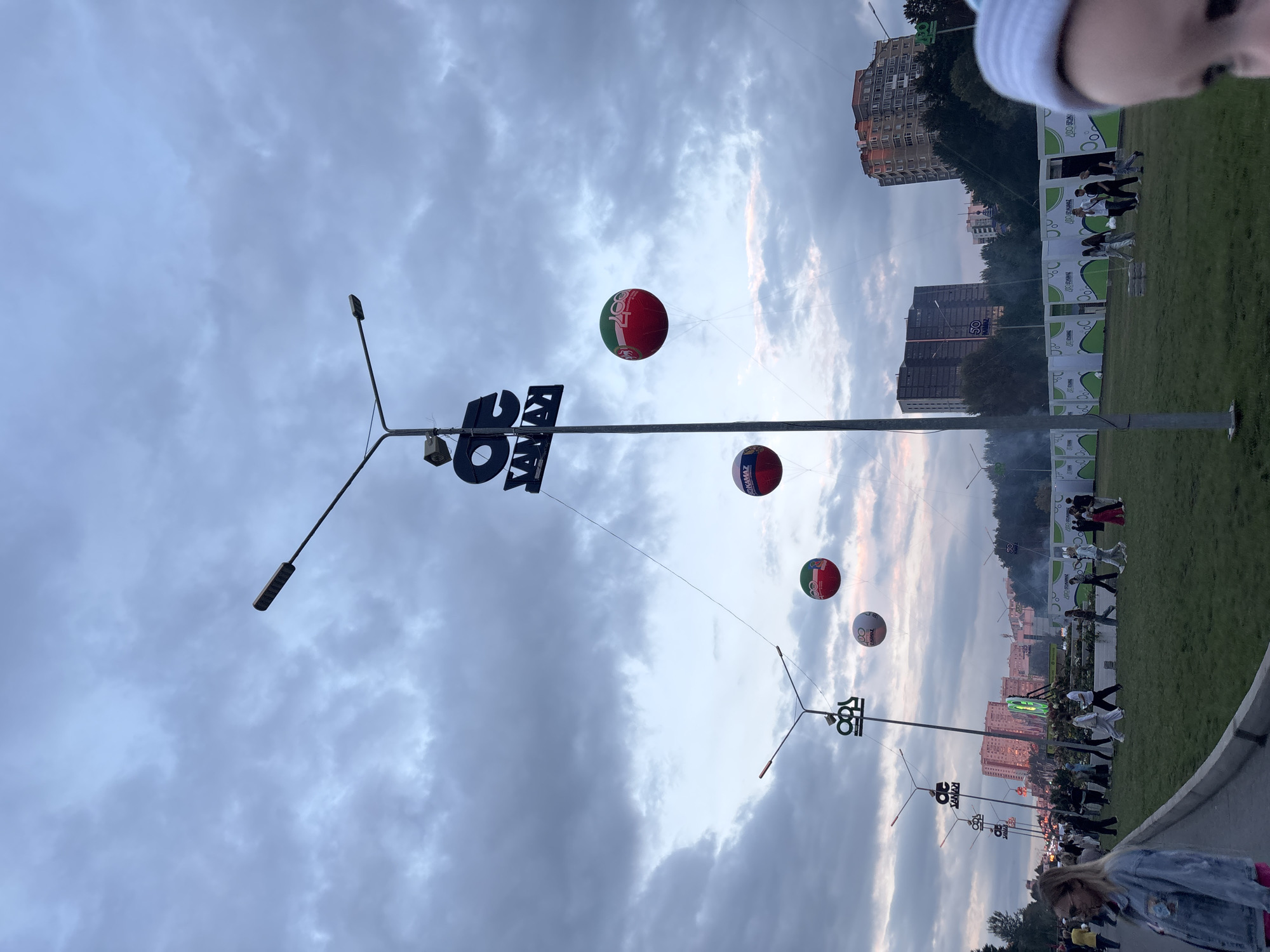 The image depicts a street scene in Naberezhnye Chelny, Russia, at dusk. A prominent lamppost with a logo is visible, and several large, spherical balloons are suspended from it by thin lines. The balloons are red and green, with some displaying logos and text. In the background, there are tall buildings, trees, and a crowd of people walking on a grassy area. The sky is cloudy and overcast, with hints of pink and orange from the setting sun. The overall atmosphere is one of an outdoor event or festival.