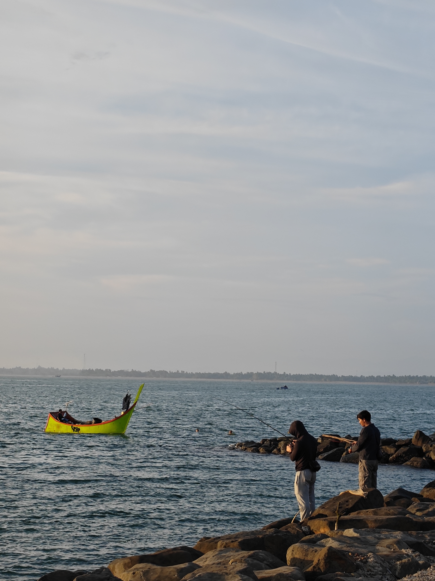 Two people stand on large rocks, fishing. One person is wearing a hooded sweatshirt and light-colored pants. The other is wearing a dark sweatshirt and pants. Both hold fishing rods. A bright yellow and green boat sits in the water. A person is visible inside the boat. The location is the city of Lhokseumawe, Indonesia. In the background, there is a landmass with trees and a communication tower. The sky is overcast. The water is choppy.