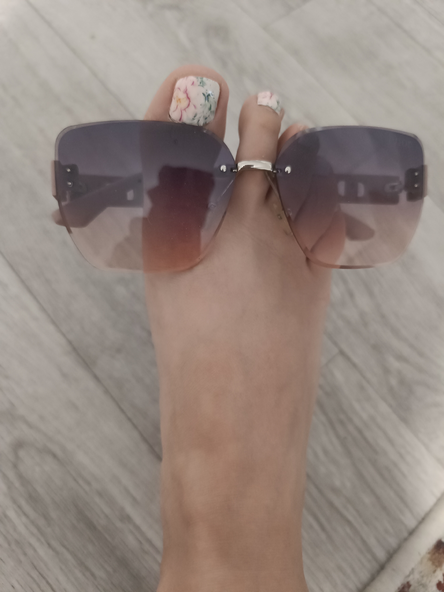 A close-up, overhead shot shows a person's foot, with painted toenails, holding up a pair of rimless sunglasses. The sunglasses have purple lenses that fade to a reddish-brown at the bottom. The foot is positioned on a light-colored wooden floor. The scene is indoors, with no specific location in Taraz city administration, Kazakhstan, identifiable.