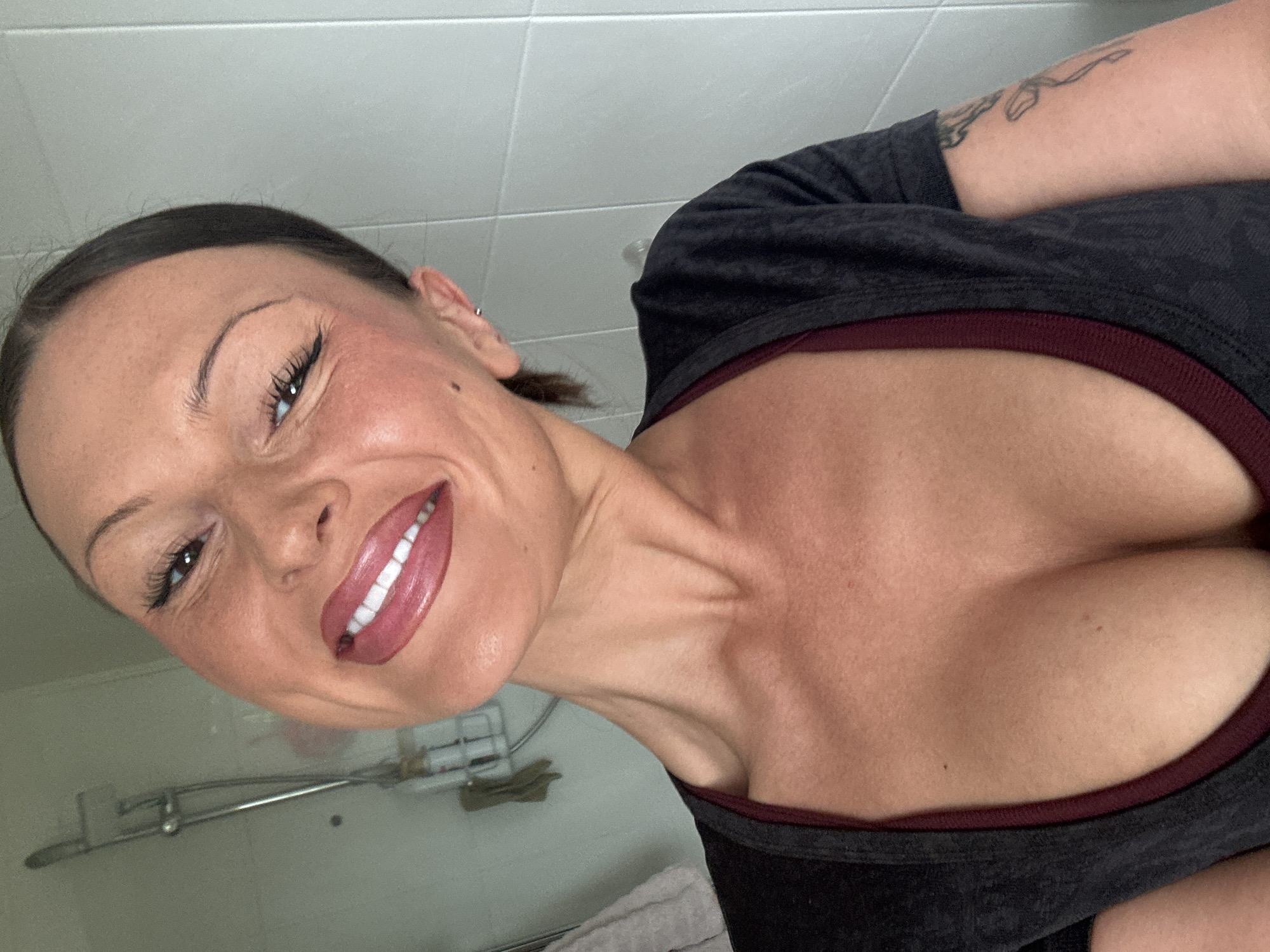 A selfie captures a woman with fair skin, brown hair pulled back, smiling with glossy pink lips. She's wearing a low-cut gray top with a maroon trim, revealing cleavage. A tattoo is visible on her upper arm. The background suggests a bathroom with white tiled walls. A chrome shower fixture is partly visible, along with a soap dish and towel. The location cannot be determined as Tunbridge Wells, United Kingdom, based on this image alone.
