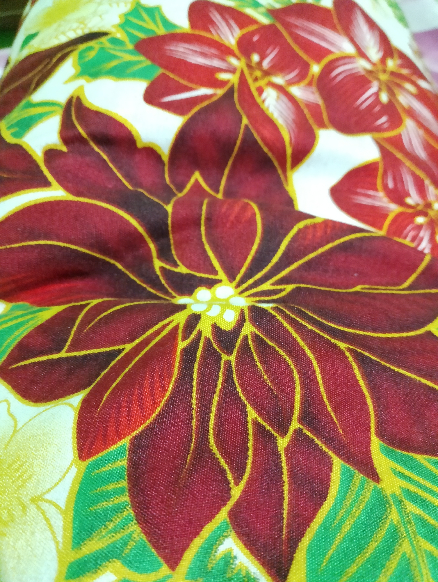 A close-up shot showcases a fabric with a floral pattern, likely a pillowcase or tablecloth, featuring large, stylized poinsettias in deep red with vibrant yellow outlines. Green leaves are interspersed between the flowers. The fabric exhibits a subtle texture from the weave. No people, actions, or specific location in Rembang, Indonesia, are identifiable in this image.