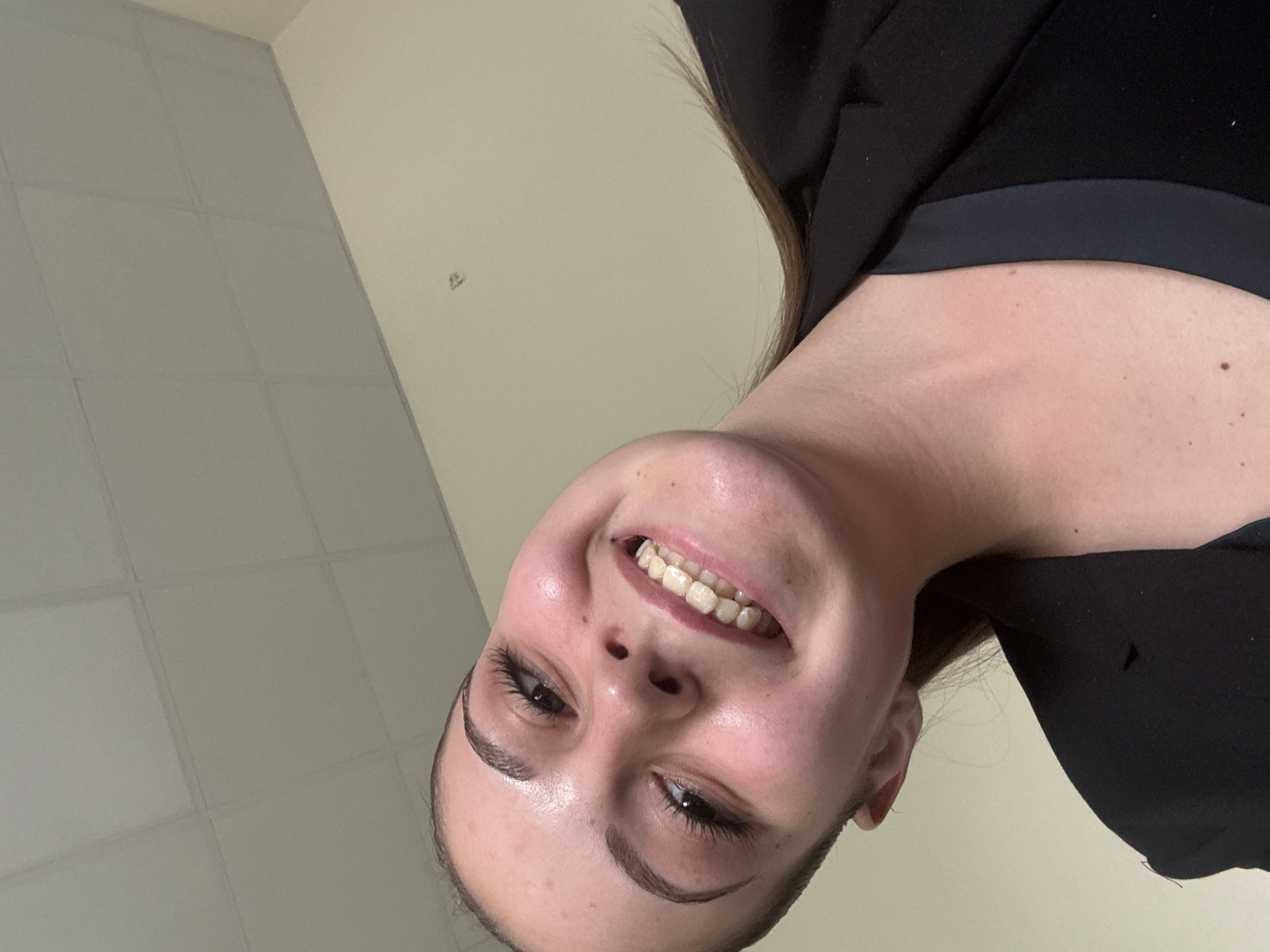 An indoor, low-angle portrait captures a young woman smiling brightly, with the image appearing rotated, placing her head upside down. The woman, who has fair skin, is seen from the chest up, wearing a dark, possibly black, top or jacket. Her light brown hair is visible along her shoulders. She is looking slightly upwards and to the side, with a wide, open-mouthed smile revealing her natural, slightly yellowish teeth. Her eyebrows are well-defined, and her eyes, with visible eyelashes enhanced by mascara, convey cheerfulness. Small freckles or moles are visible on her neck and upper chest.
The background primarily consists of a plain, light-colored wall, likely cream or off-white, and a section of a greyish drop ceiling with visible square panels. A small, indistinct mark or fixture is present on the wall. The lighting appears to be even, suggesting indoor artificial light. The overall mood conveyed by her expression is one of happiness or amusement. There is no visible text in the image. The scene is located indoors within Burgas, Bulgaria.