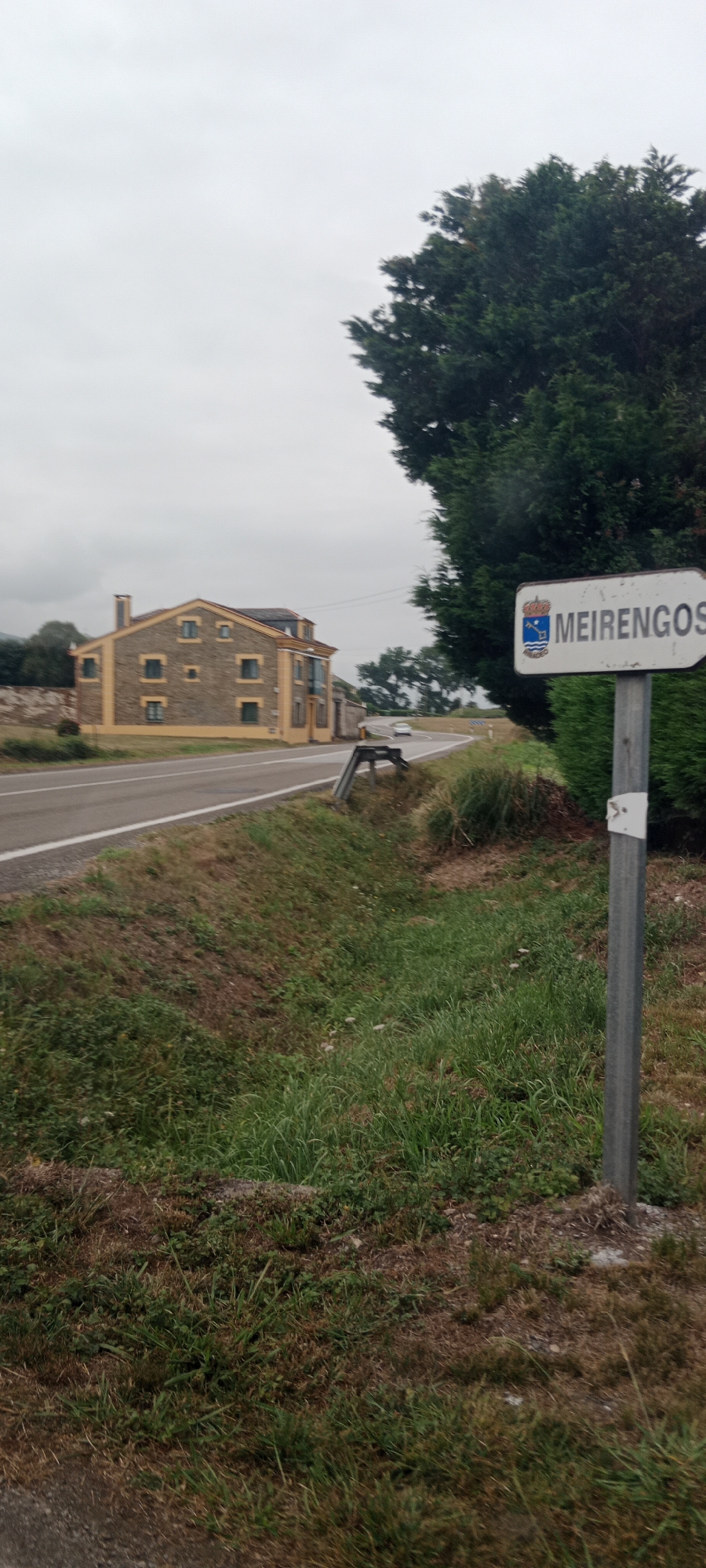 The image shows a signpost for "MEIENGOS" located in Zopetal, Spain, on a cloudy day. The signpost is white with blue and red lettering and features a coat of arms. It stands on a metal pole next to a dense green hedge. To the left of the signpost, a road curves away into the distance. Beside the road, a two-story house with a yellow facade and brown stone accents is visible. The house has multiple windows and a chimney. The surrounding environment consists of grassy slopes and trees, with one large, dark green tree dominating the right side of the frame. A white car is seen driving away on the road. The overall impression is of a rural or semi-rural setting.