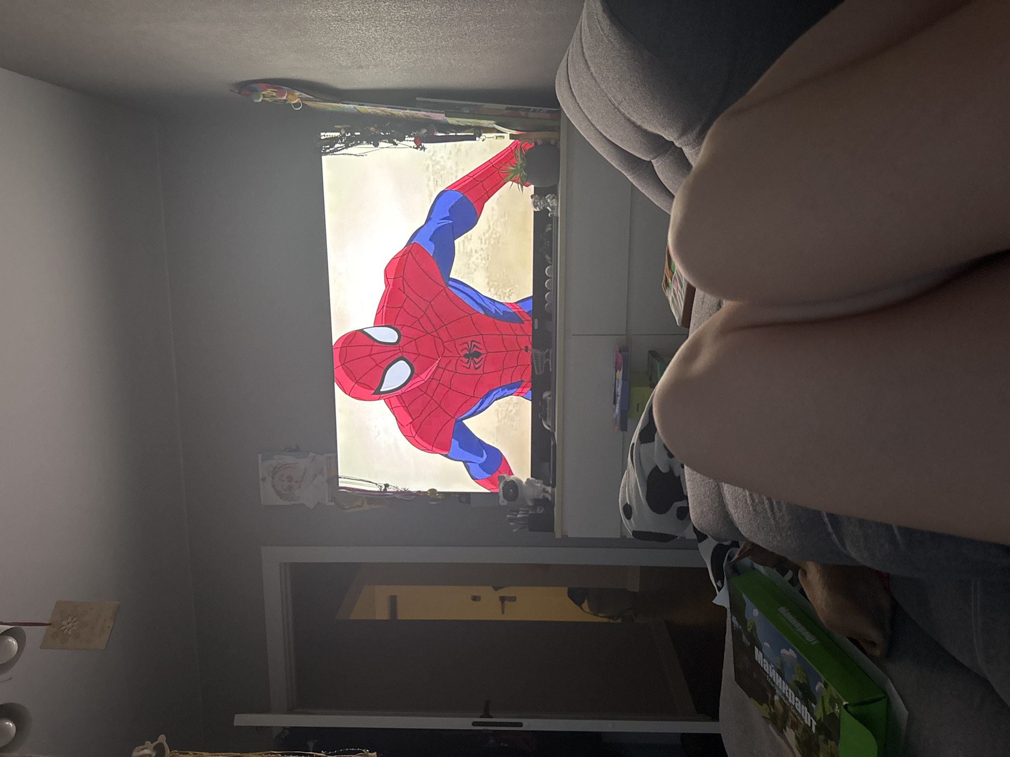 The image shows a person sitting on a grey sofa, watching a television. The television is displaying an animated image of Spiderman. The setting seems to be a room with a door leading to another room. Various objects and items are visible on top of and around the television stand, including a Minecraft box. The exact location within Chaikovsky, Russia cannot be determined from the image.