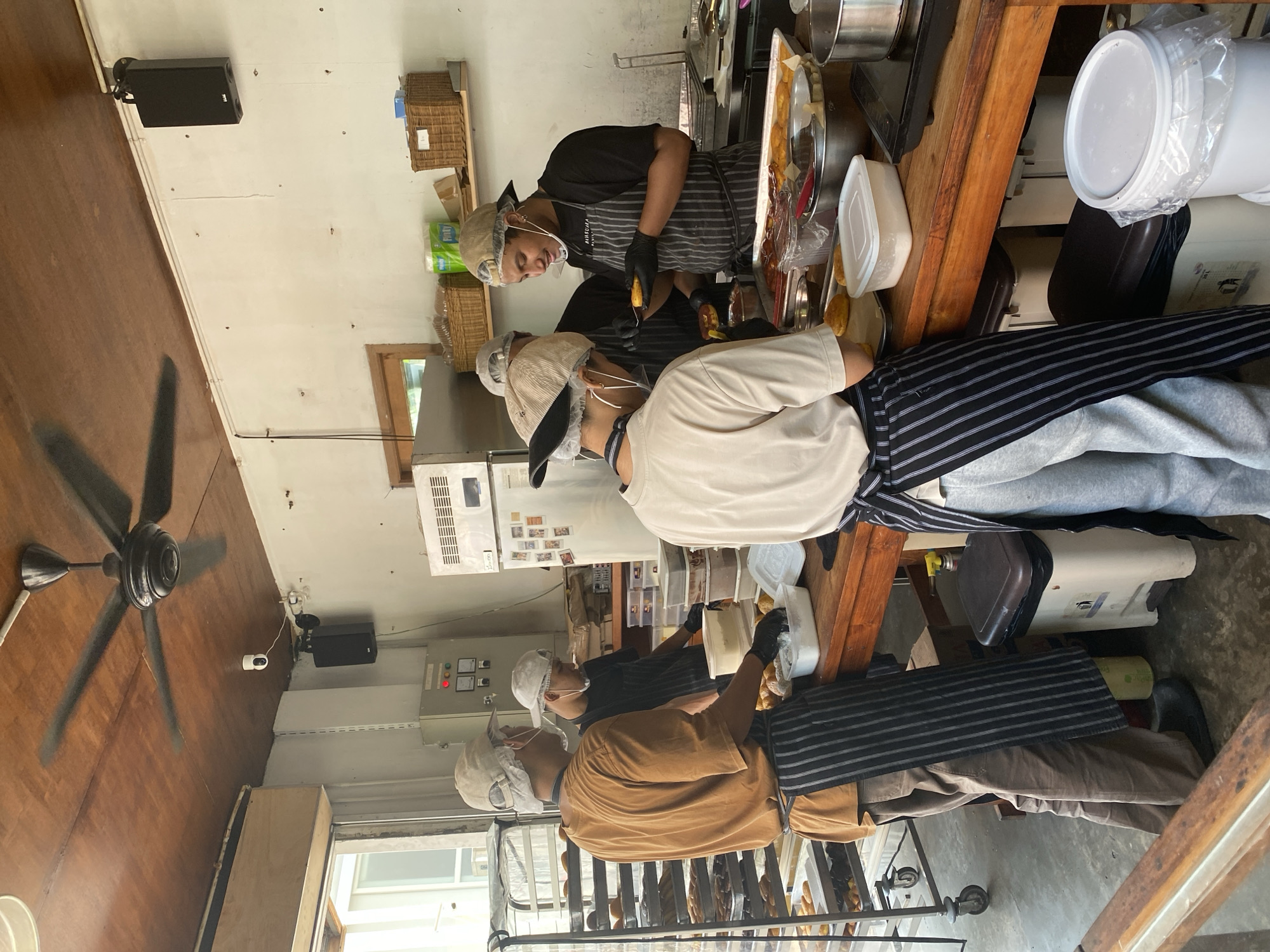 In a brightly lit commercial kitchen, likely in South Jakarta, Indonesia, four individuals are engaged in food preparation. Two men, wearing chef's hats and aprons, stand at a large wooden workbench. One, in a brown t-shirt and a black and white striped apron, faces away from the camera and appears to be handling food items on a tray. The other, in a black t-shirt and a similar apron, faces the camera and holds a yellow, pastry-like item, seemingly in the process of placing it into a container. A third person, also in a chef's hat and apron, stands between them, looking down at the work. A fourth individual, in a light-colored t-shirt and an apron, is partially visible at the front of the workbench, also attending to food items. The environment includes stainless steel appliances, shelving units filled with baked goods, and various food containers. A ceiling fan circulates air above the scene. The overall impression is of an active, bustling food production environment.