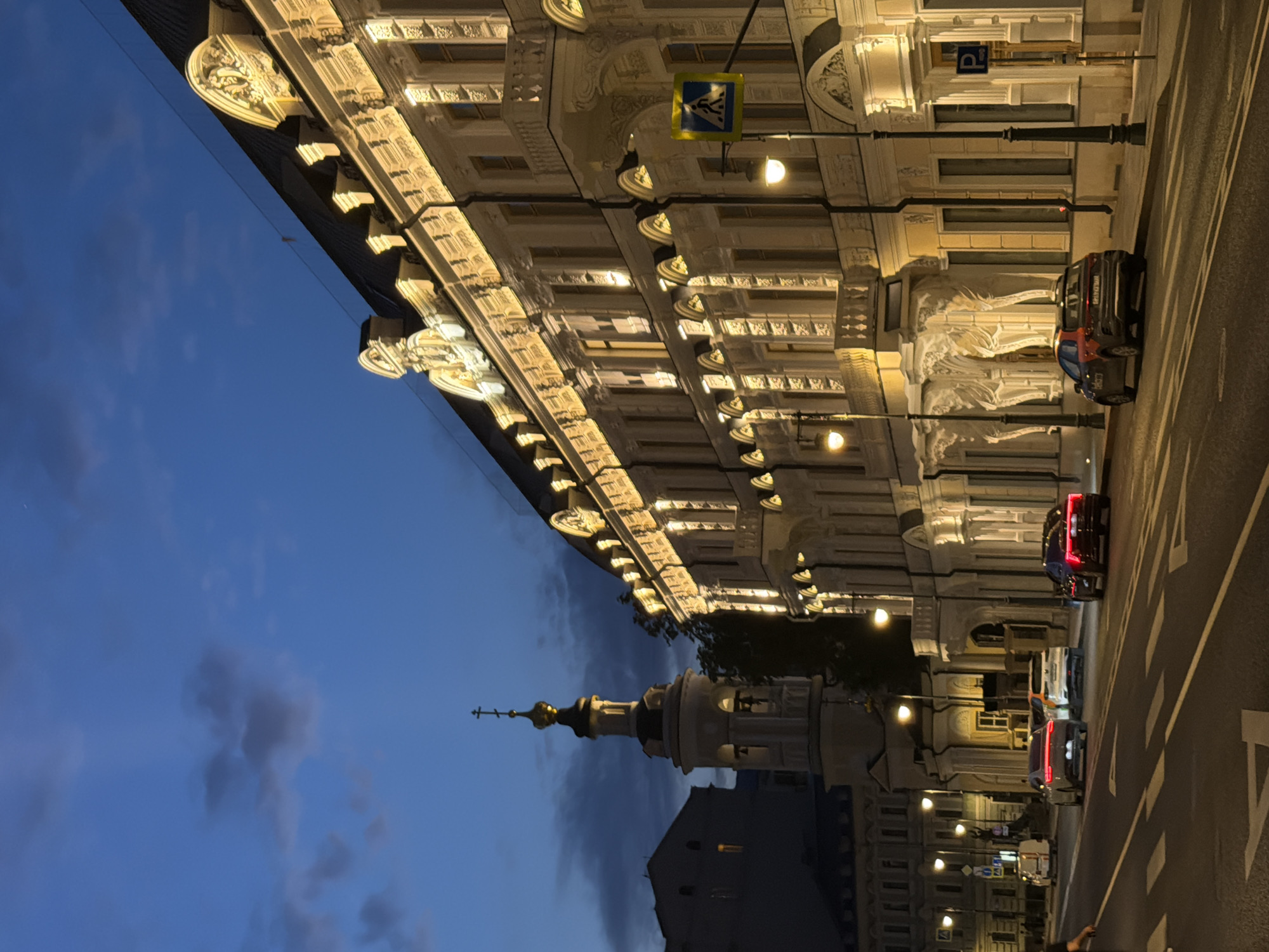 The image captures a nighttime view of a street in Moscow, Russia, likely in the area of Patriarch's Ponds or a similar historic district. The scene is dominated by a grand, ornate building illuminated with warm artificial light, highlighting its classical architecture, balconies, and decorative elements. Across the street, a bell tower with a golden dome and a cross stands against the twilight sky, suggesting the presence of a church or monastery nearby. Several cars are visible on the asphalt road, with their headlights and taillights illuminated. One car, a dark sedan, is parked along the sidewalk in the foreground. The sky is a deep blue, with some scattered clouds, indicating the transition from dusk to night. Streetlights and illuminated building windows contribute to the overall ambiance. A pedestrian crossing sign is also visible, denoting a marked crosswalk on the road. The image appears to be a candid street photograph, focusing on the interplay of architecture, light, and urban life at night.