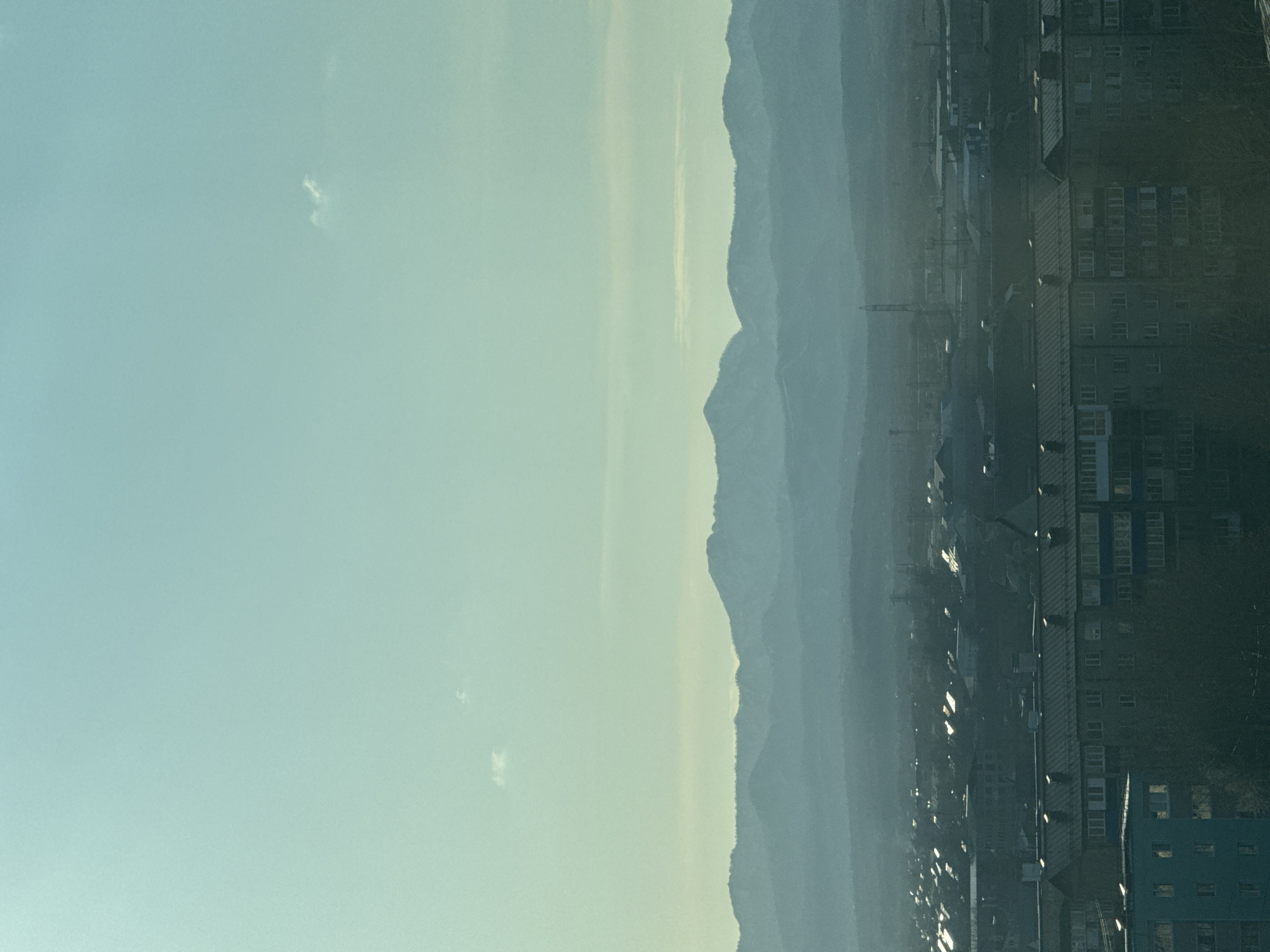 An atmospheric landscape photo captured from an elevated perspective shows a blend of urban and natural elements under a hazy sky. The image, when correctly oriented, features a pale blue-green sky at the top, transitioning into a lighter, almost yellowish hue near the horizon, indicative of atmospheric haze or a low sun angle. A few small, wispy white clouds are scattered across the sky.
In the middle ground, a prominent mountain range stretches across the scene, appearing in multiple layers of hazy blue and grey. The nearest mountain silhouettes are more defined, showing rugged peaks, while distant ranges are softened by the atmospheric conditions.
The foreground and midground on the right side are occupied by a cityscape. Dark, multi-story buildings with numerous windows and some visible balconies dominate this section. Some windows reflect a bright, diffused light, suggesting the time of day is either early morning or late afternoon. To the left, further into the city, hints of industrial structures, including what appears to be a crane, can be discerned amidst the haze.
No people are visible. No specific activity or event is clearly taking place. The overall impression is one of quiet vastness, with the city nestled at the foot of towering, misty mountains. No visible text is present in the image. The scene is consistent with views found in mountainous regions near urban centers, such as Yuzhno-Sakhalinsk, Russia.