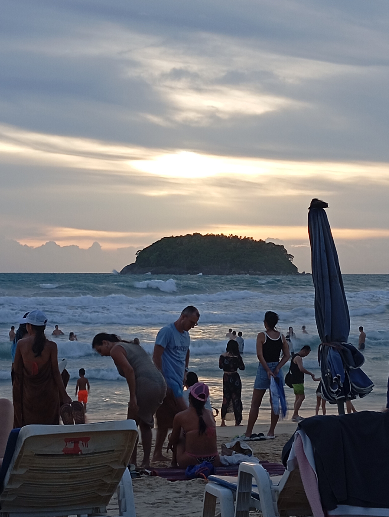 This is a photograph taken at Karon Beach, Thailand, during sunset. The sky is filled with dramatic clouds, tinged with orange and yellow from the setting sun, which is partially obscured by the clouds. A lush green island with a tree-covered peak stands in the middle distance on the ocean. The sea is wavy, with white foam cresting on the waves as they approach the shore.
On the beach, several people are visible. In the foreground, a beach chair is partially in frame, with a towel draped over it. To the left, a woman wearing a white hat and a brown wrap stands with her back to the camera. Next to her, a woman in a grey dress bends over. Further out in the water, several people are wading and swimming, silhouetted against the waves.
To the right of the frame, a large, dark blue beach umbrella is folded and leaning precariously, with towels and other items attached. Several people are gathered near the umbrella, including a woman in a black tank top and denim shorts, and a man in a black tank top and green shorts. A child in orange shorts is also visible in the water, walking towards the shore. The overall scene depicts a relaxed atmosphere at a popular tourist beach as the day concludes.