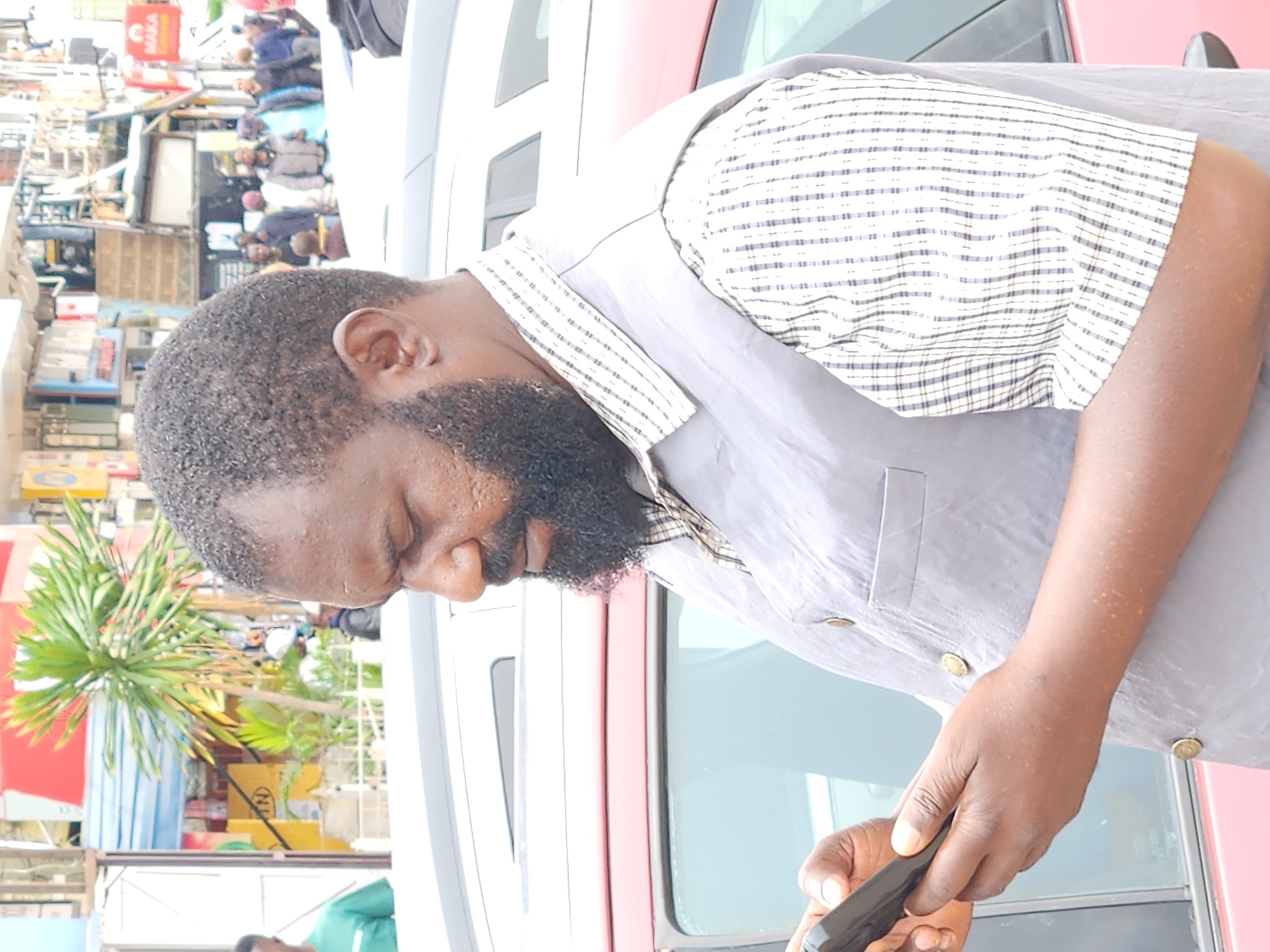 The image captures a medium shot of a Black man with a beard and short hair, wearing a light grey vest over a checkered shirt. He is holding and looking at a black smartphone in his hands. The man is positioned in front of a red vehicle, with another white vehicle partially visible in the background. The environment appears to be a city street, with buildings and other people visible in the distance. The background suggests an urban setting, possibly a marketplace or a busy street with shops and vehicles.