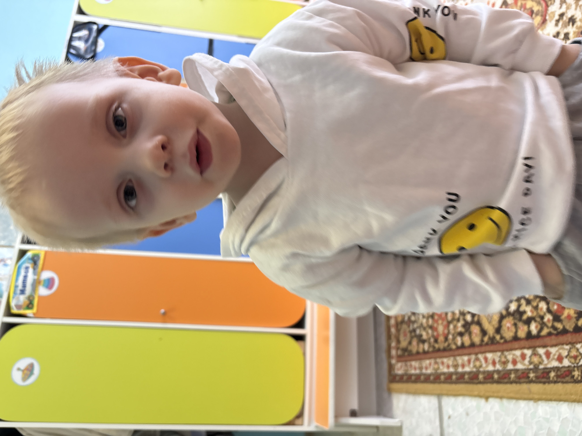 The image captures a young child standing indoors within what appears to be a childcare facility in the city of Chaikovsky, Russia. The child, with light blonde hair and light-colored eyes, is centered in the frame, looking directly at the camera. They are wearing a white hooded sweatshirt adorned with yellow smiley face graphics and partial text, including "SMILE YOU" and "NICE DAY!" on the torso, and a similar graphic on the left sleeve. The child's hands are placed inside the pockets of their visible grey pants. In the background, a row of multi-colored storage cubbies or lockers is present: a lime green unit, an orange unit with a small silver handle and a rectangular label displaying "Малыши" (Malyshi) along with child-related imagery, and a blue unit, all featuring rounded top edges. A light-colored bench or shelf runs beneath these units. The floor in the foreground is covered by a patterned rug with dark, red, and gold floral designs, adjacent to a lighter, possibly tiled, floor section on the far left.