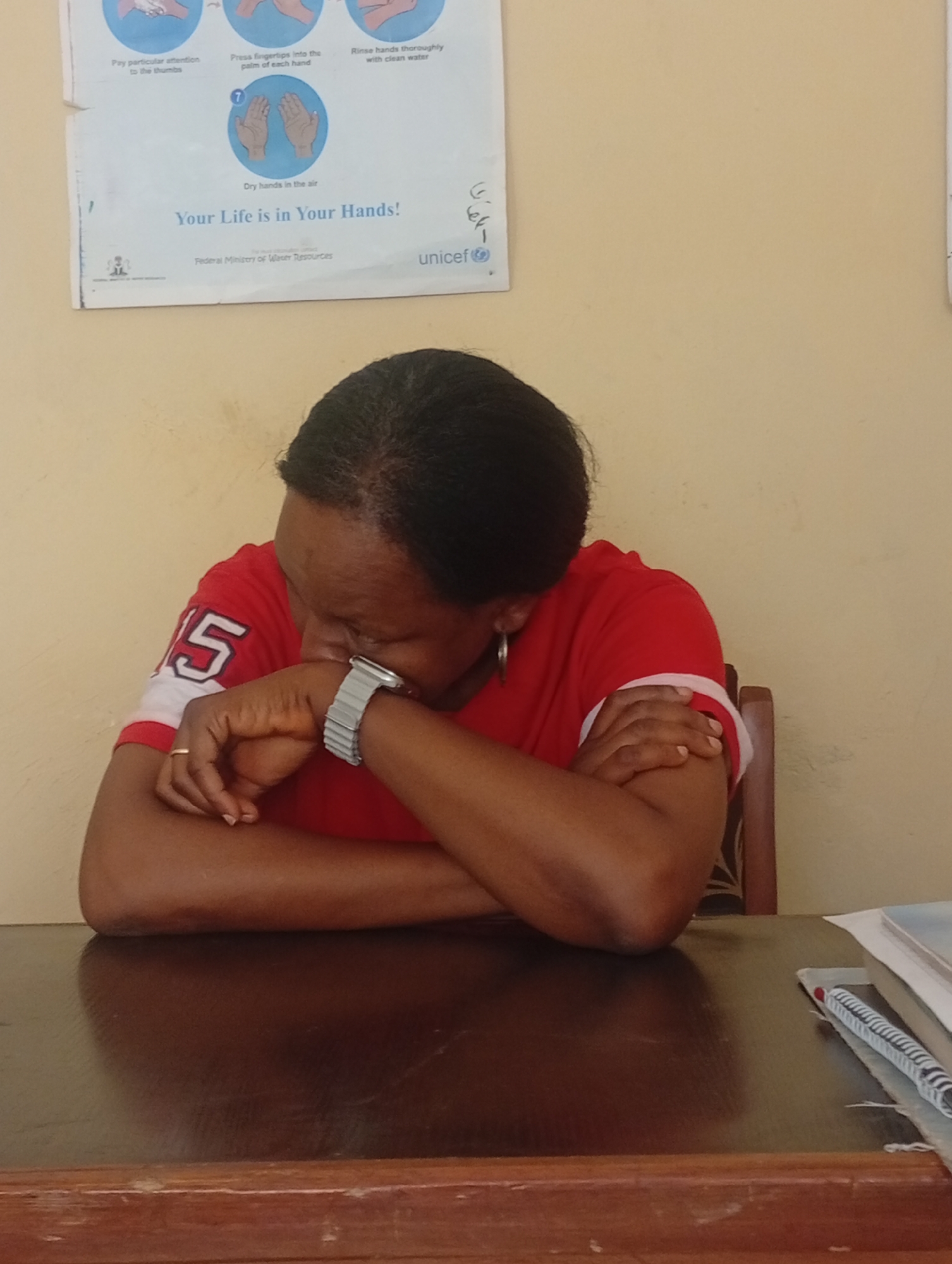 An indoor, eye-level shot captures a woman seated at a dark wooden table, her head resting on her crossed arms. She appears to be either upset, exhausted, or deeply contemplative. The woman has dark skin and black hair pulled back. She wears a red t-shirt with a white trim and the number "15" visible on the right sleeve. A silver-toned watch is on her left wrist, and a simple gold-colored ring is on her left ring finger. A hoop earring is visible on her left ear.
Behind her, against a plain, light yellow wall, hangs a poster illustrating proper hand-washing techniques. The poster features several small diagrams depicting steps like "Pay particular attention to the thumbs," "Press fingertips into the palm of each hand," "Rinse hands thoroughly with clean water," and "Dry hands in the air." A prominent tagline reads, "Your Life is in Your Hands!" Below this, text identifies "Federal Ministry of Water Resources" and the "unicef" logo.
To the right of the woman on the table, a stack of spiral-bound notebooks or books and a red-capped pen are visible. The setting, an unknown indoor location, possibly a public office or educational facility, is subtly indicated by the informative poster. The lighting suggests daytime indoors, but the exact time is indeterminate. The overall mood is somber due to the woman's posture and apparent emotional state.