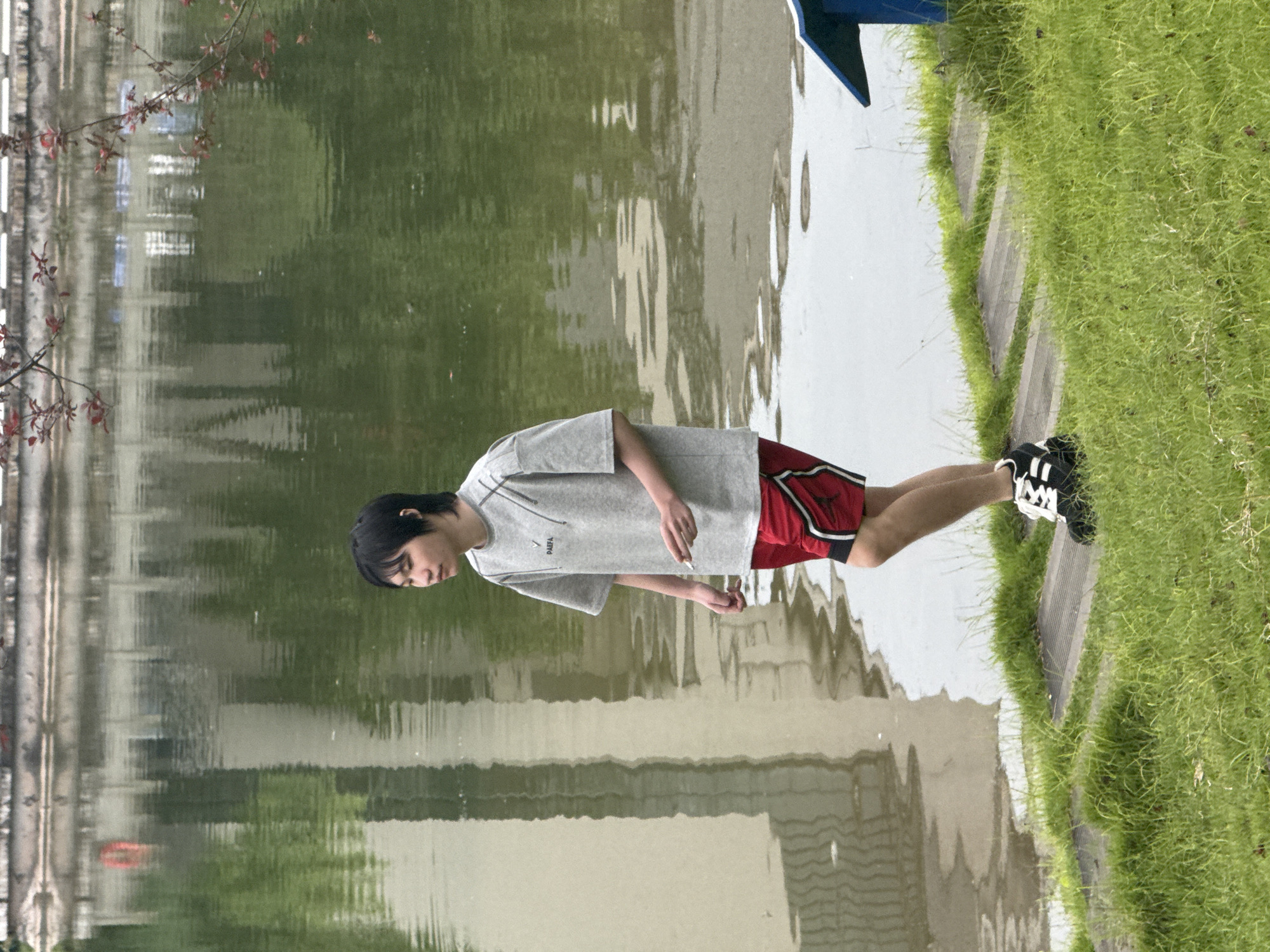 A young person with dark hair stands beside a body of water in what appears to be a park or recreational area. They are wearing a grey t-shirt with some dark lines and text that is partially obscured but reads "DAPPA", red shorts with black trim, and black and white athletic shoes. The person is looking down towards their hand, which is holding a cigarette. The scene is dominated by the reflective surface of the water, which mirrors the surrounding environment, creating an abstract, wavy pattern. To the right of the water, a neatly mown grassy bank slopes upwards, with a concrete path or edge visible beneath the grass. In the background, a tree with small red leaves can be seen on the left, and some blurred structures are reflected in the water. A small portion of a blue and white object, possibly part of a boat or structure, is visible at the top right. The lighting suggests it is daytime, and the presence of foliage indicates a season other than winter. The overall mood appears contemplative.