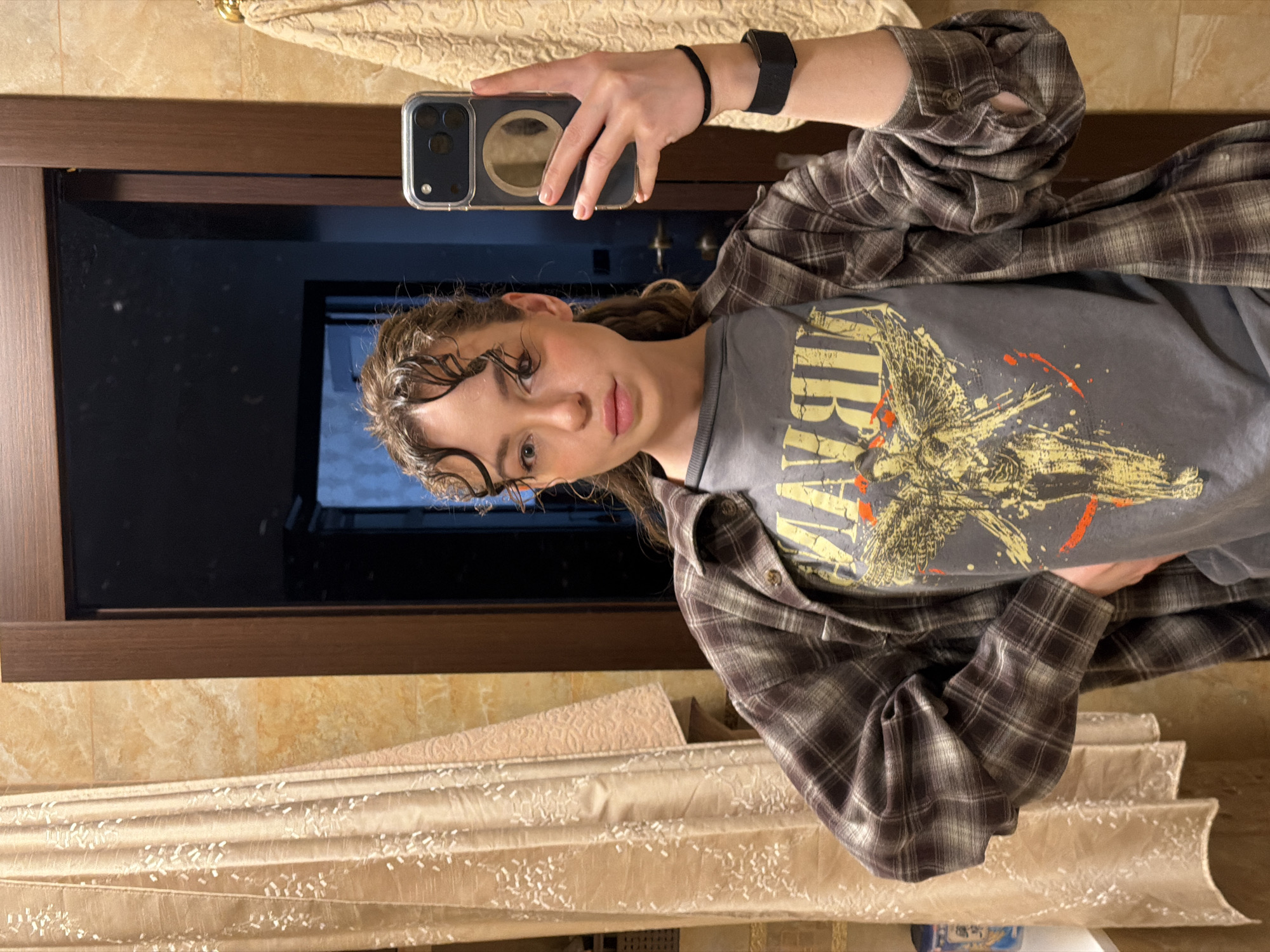 A person is taking a selfie in a bathroom. The person has brown, curly hair that is wet and slicked back. The person is wearing a gray graphic t-shirt, a brown and white plaid flannel shirt, and a black smart watch on their left wrist. They are holding a phone with a clear case in their right hand. The background consists of a mirror reflecting a dark room, a beige tiled wall, and a beige and white patterned curtain. The location is not identifiable as Khimki, Russia, based on the image provided.