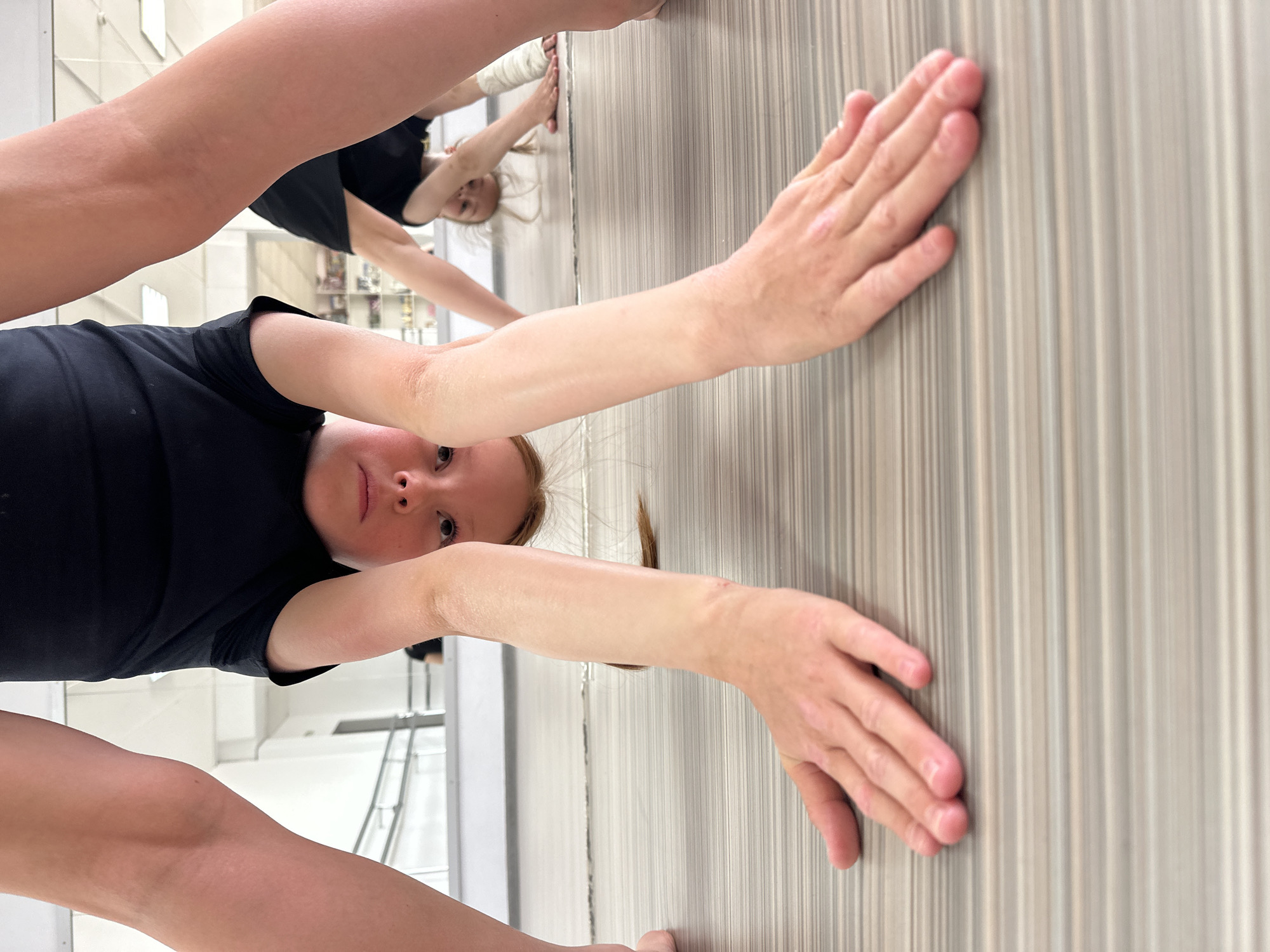 A low-angle shot shows a young, light-skinned, red-haired girl wearing a black t-shirt in what appears to be a dance studio. Her arms are extended forward, with her hands pressed against a striped wall. A partial view of another person's arm and leg is in the foreground, suggesting a dance instructor or fellow student. A reflection in a mirror shows another girl with blonde hair. The walls of the studio are white, and there is a barre in the background.
Because the image was provided with the information that the location is Votkinsk, Russia, it would be a dance studio located in that city.
