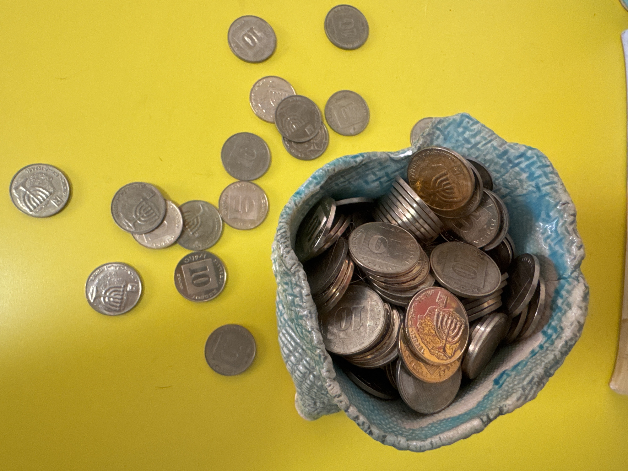 The image shows a collection of Israeli coins, specifically 10 agorot pieces, both inside and scattered around a small, light blue ceramic bowl. The coins are mostly silver in color, with some displaying a prominent "10" and others featuring a Menorah emblem. The bowl is placed on a yellow surface. There is no definitive indication of the location within Ramat Gan, Israel, in the image.