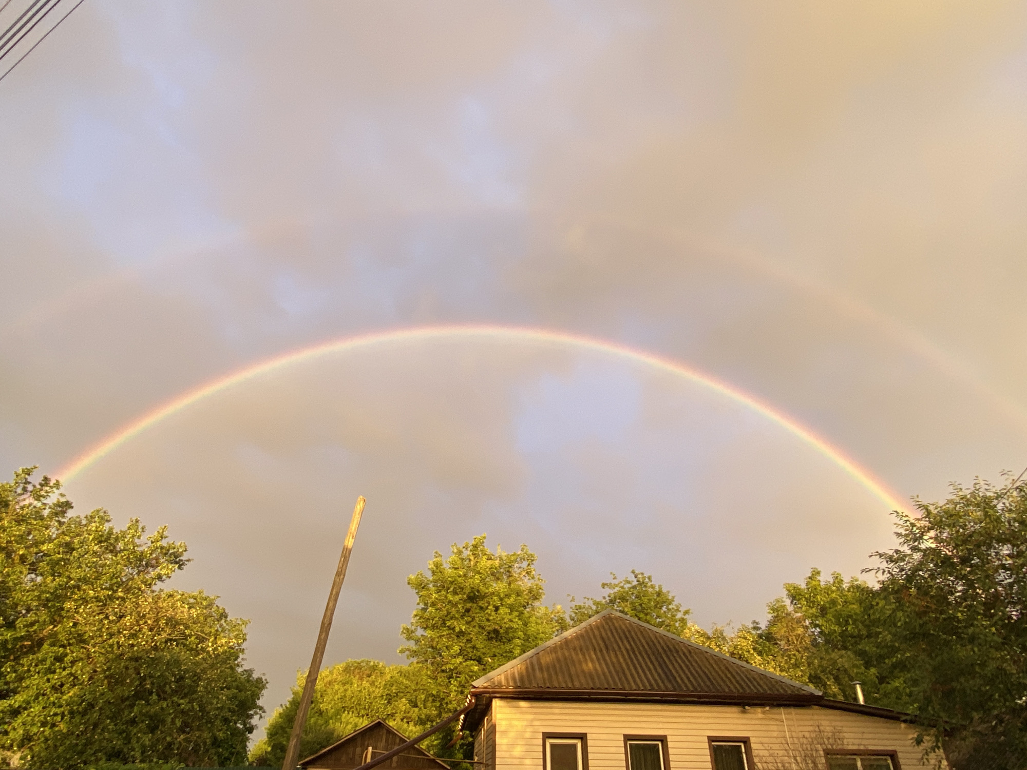 The image captures a vibrant double rainbow arcing across a pale, cloudy sky. The sky transitions from a light grey on the left to a warm, golden hue on the right. Below the rainbow, a residential scene unfolds. Lush green trees, illuminated by the soft light, frame the view on both sides. In the foreground, a light-colored house with a corrugated metal roof and dark-trimmed windows is visible, nestled among the trees. To the left of the house, another smaller, dark-roofed structure can be seen. A weathered wooden pole stands prominently near the center of the image, adding a rustic element to the composition. The overall atmosphere is serene and picturesque, suggesting a peaceful rural or suburban setting.