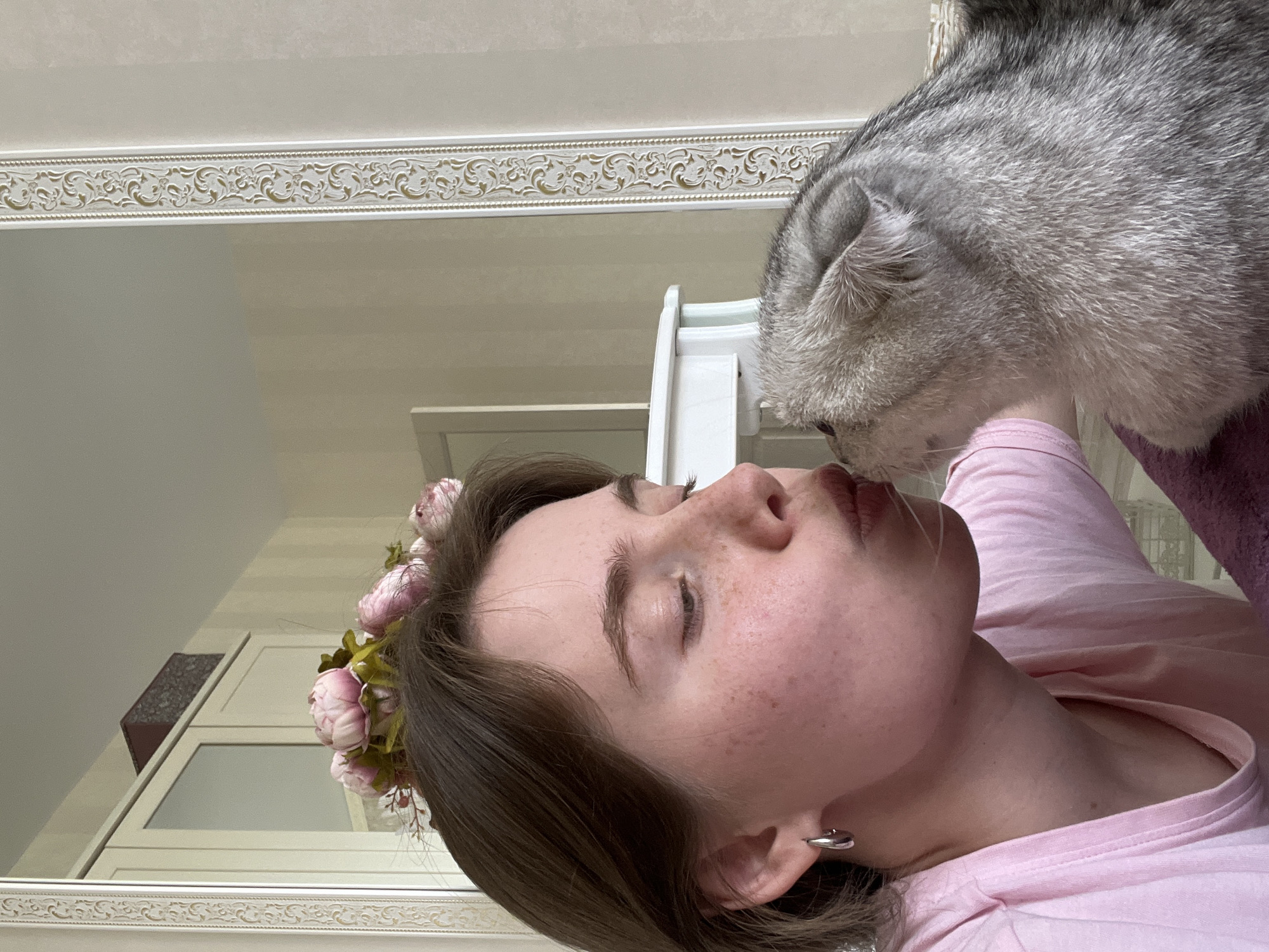 A young woman with fair skin, freckles, and short brown hair adorned with a floral crown lies on her back, wearing a pink top. Her eyes are closed. A large, gray tabby cat is positioned near her face, appearing to nuzzle or sniff her. The scene appears to be indoors, possibly in a bedroom, with a white-framed mirror behind them. The mirror reflects a portion of the room, including a white dresser or cabinet. A decorative border is visible at the top of the mirror. The location is Chaikovsky, Russia.