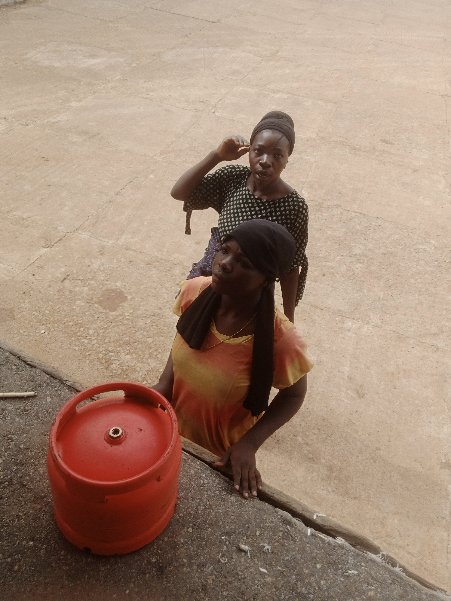 Two young women are standing outdoors on a paved surface. In the foreground, a red gas cylinder is placed on a raised concrete ledge. The woman closer to the cylinder is wearing an orange and yellow ombre t-shirt and a black headscarf. The second woman, standing behind her, is wearing a black and white patterned blouse and a black headscarf. She is looking towards the camera with her hand raised to her ear. The ground appears to be concrete with visible seams and some debris. The overall impression is a scene likely in Jos, Nigeria, given the context provided.