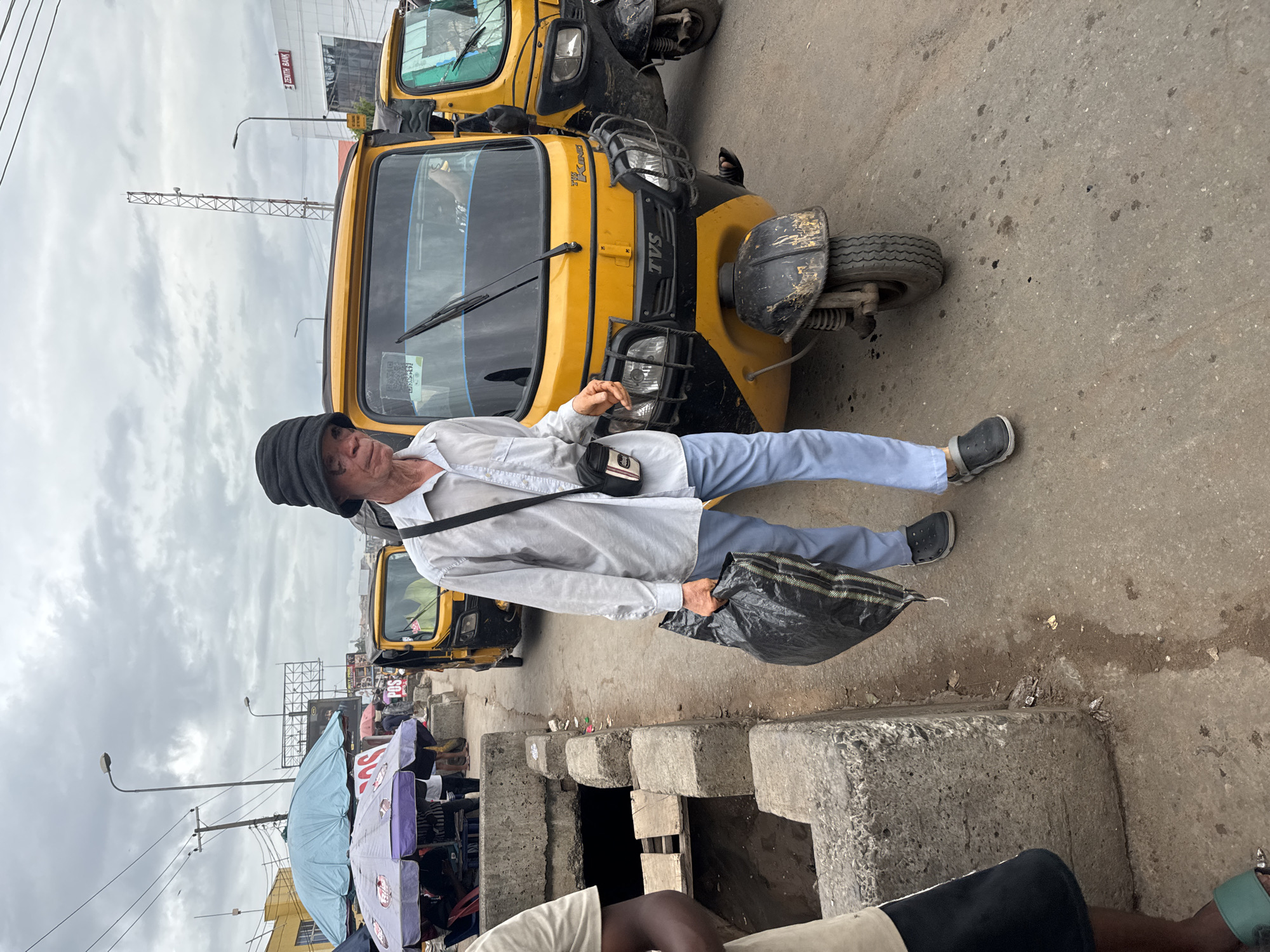 The image depicts a street scene in Nkpor, Nigeria, under an overcast sky. A man wearing a white shirt, light blue pants, black shoes, and a dark hat is standing on the roadside, holding a black plastic bag. He has a small bag slung across his shoulder. He is standing near a yellow "keke", a type of auto-rickshaw, which is parked on the street. Several other kekes are visible in the background. To the left, a concrete barrier separates the road from a lower area where people and vendors with umbrellas are situated. The street surface is cracked and shows signs of wear. A communication tower is visible in the background.