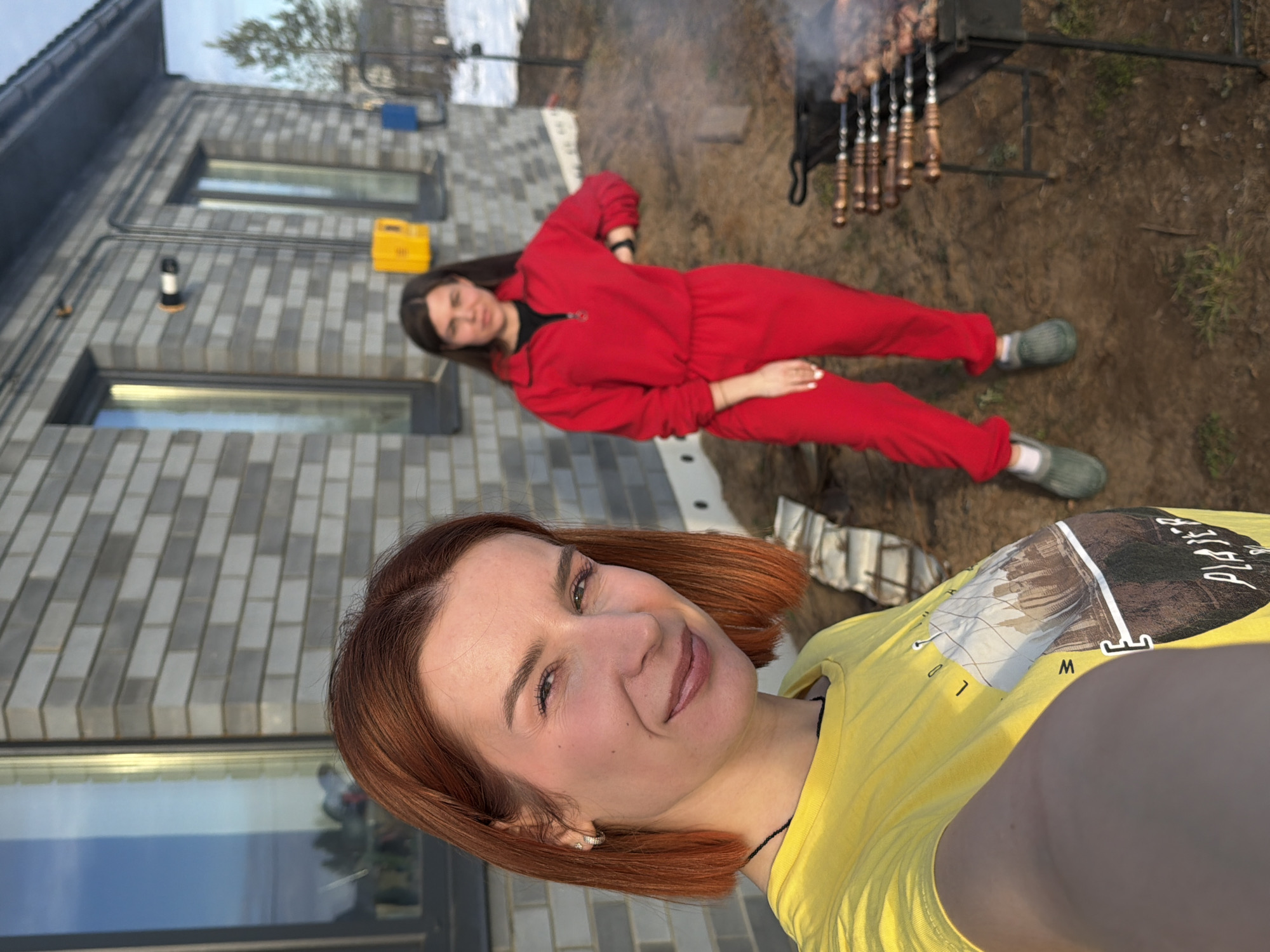 The image depicts an outdoor scene in Chaikovsky, Russia, featuring two adult females and a barbecue setup in front of a modern brick house.
In the foreground, a female with short, reddish-orange hair is positioned to the left, taking a selfie. She is smiling and looking directly at the camera. She wears a yellow t-shirt with a partial graphic featuring a cityscape and the visible text "PLAYER." A small silver hoop earring is visible in her left ear, and a dark necklace is worn around her neck.
In the mid-ground, a second female stands slightly to the right of the frame, partially obscured by the foreground subject. She has long, dark hair and wears a red, full-body jumpsuit or tracksuit with a black half-zip collar. Her left hand is placed on her hip, and she wears green or grey clogs. She is looking towards the camera.
Between the background female and the right edge of the frame, a black metal barbecue brazier (mangal) is present, holding several skewers (shashlik) of meat cooking over visible coals, emitting smoke.
The background features a two-story house with a grey brick facade and dark-framed windows. A light fixture and a yellow electrical box are mounted on the exterior wall. Patches of snow and bare earth with sparse vegetation are visible on the ground. A pine tree is partially visible to the far right. The scene is illuminated by natural daylight.