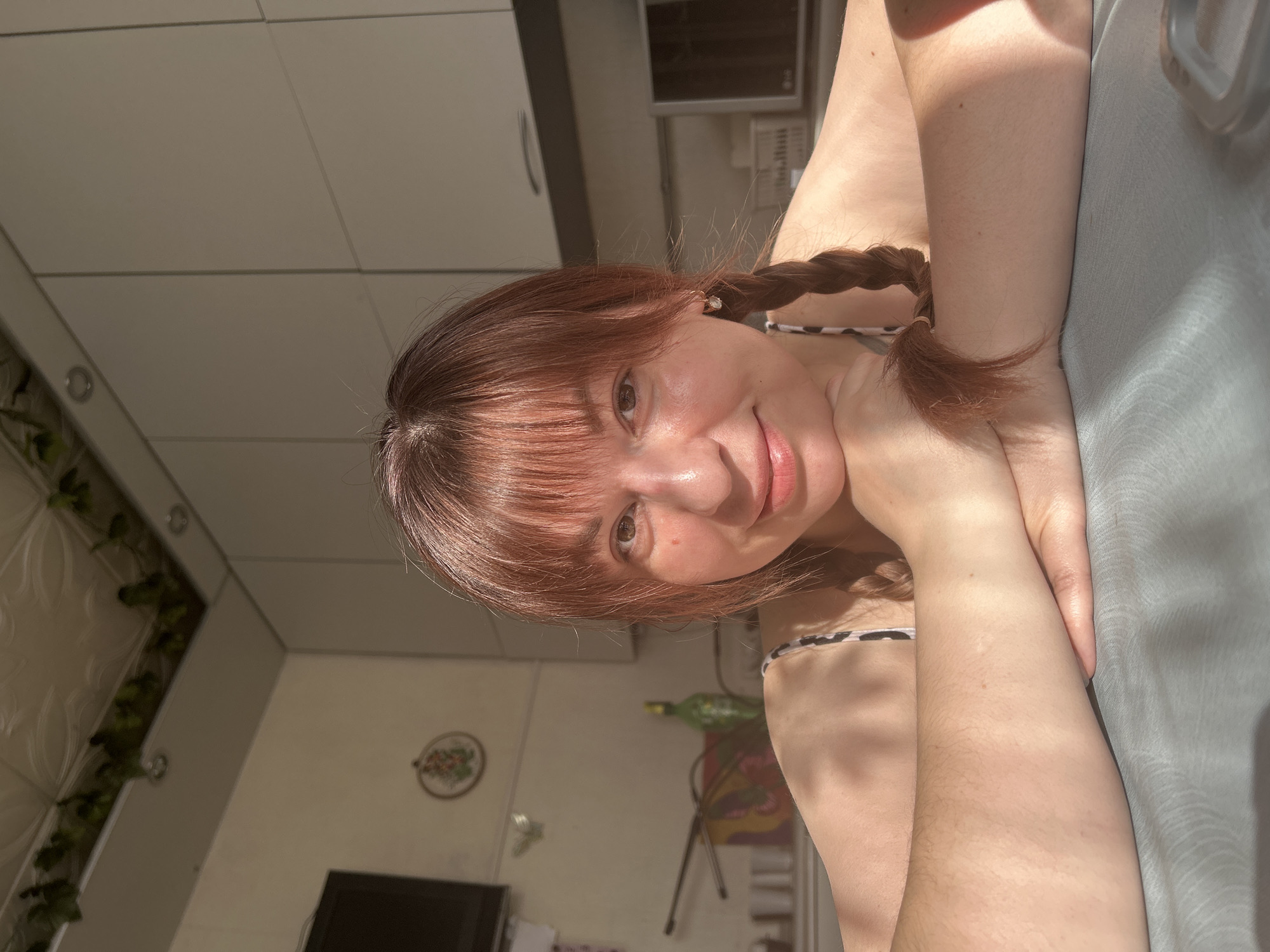 The image is a brightly lit, close-up portrait of a young woman taken indoors, likely a selfie or a photograph from a close vantage point. The woman has shoulder-length, reddish-brown hair styled with bangs across her forehead and two braids, one visible over each shoulder. Her complexion is fair, and a small reddish spot is visible below her right eye. She is looking directly at the camera with a soft, gentle smile, showing a slight dimple on her right cheek.
She appears to be lying down or leaning back, with her right arm bent, and her hand supporting her head or resting on a light-colored surface, possibly a pillow or soft furnishing, which occupies the right side of the frame. She is wearing a light-colored top with a subtle pattern, possibly a halter top or camisole, part of which is visible around her neck and shoulder. A small, light-colored earring can be seen in her right ear.
The scene is illuminated by warm, direct sunlight coming from the upper left, casting strong highlights on her hair, skin, and the surfaces around her, creating a bright and inviting atmosphere.
The background suggests a casual home environment, possibly a kitchen or living area. To the left, white or light grey cabinet doors or wall panels are visible, with horizontal seams. Further to the left, there's a textured wall with some green, leafy artificial plants or vines hanging vertically. On the wall further in the background, a circular framed object with a floral embroidery pattern can be seen. Below the cabinets, glimpses of other household items are visible, including what appears to be a dark screen (possibly a television or monitor) and a green bottle on a surface. The ceiling, visible in the upper background, shows light-colored panels with grid lines.
No discernible text is visible in the image.