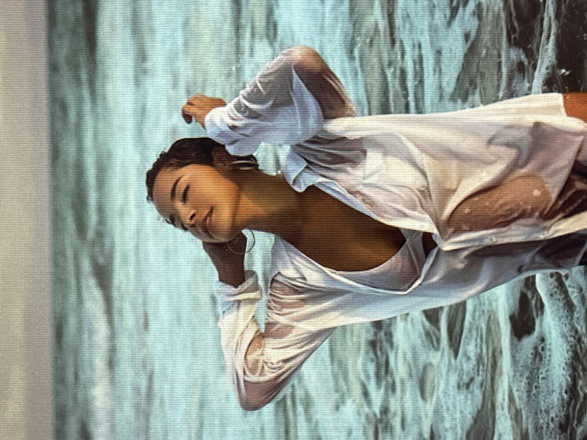 A woman is lying down in water. Her eyes are closed and her head is tilted back. She is wearing a wet white shirt that clings to her body. The water is a light blue color and appears to be moving, with some white foam on the surface. The background is blurred and appears to be a continuation of the water. The lighting suggests it might be daytime, possibly overcast. The overall mood is serene and peaceful. There is no visible text in the image.