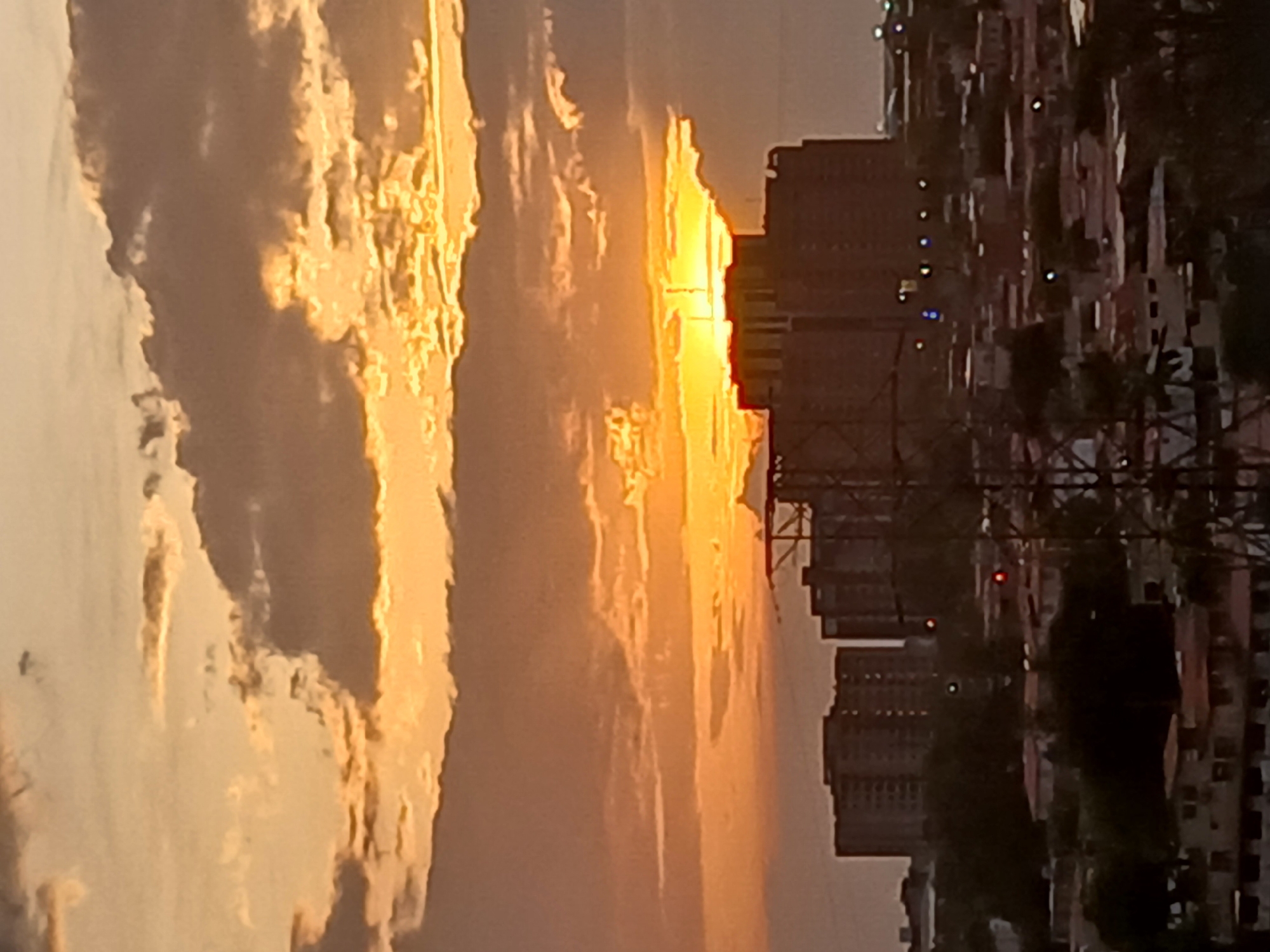 The image shows a sunset over Ciudad Guayana, Venezuela. The sky is filled with clouds that are illuminated with an orange light from the setting sun. The sun's rays break through a gap between the clouds and the city skyline. Several high-rise buildings are visible in the middle ground, with some being silhouetted. There are also power lines or a communication tower in the foreground, obscuring the cityscape. In the background, there are other buildings and trees.