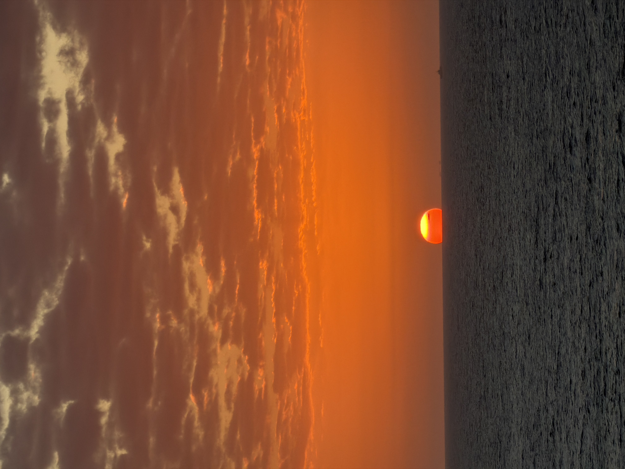 Here is a description of the image:
The image shows a landscape scene at sunset. The sun, appearing as a bright orange orb, is positioned near the horizon. To the left of the sun, the sky is filled with layers of clouds, bathed in orange and yellow hues. To the right of the sun, a dark, textured surface extends into the foreground, possibly representing land or water.