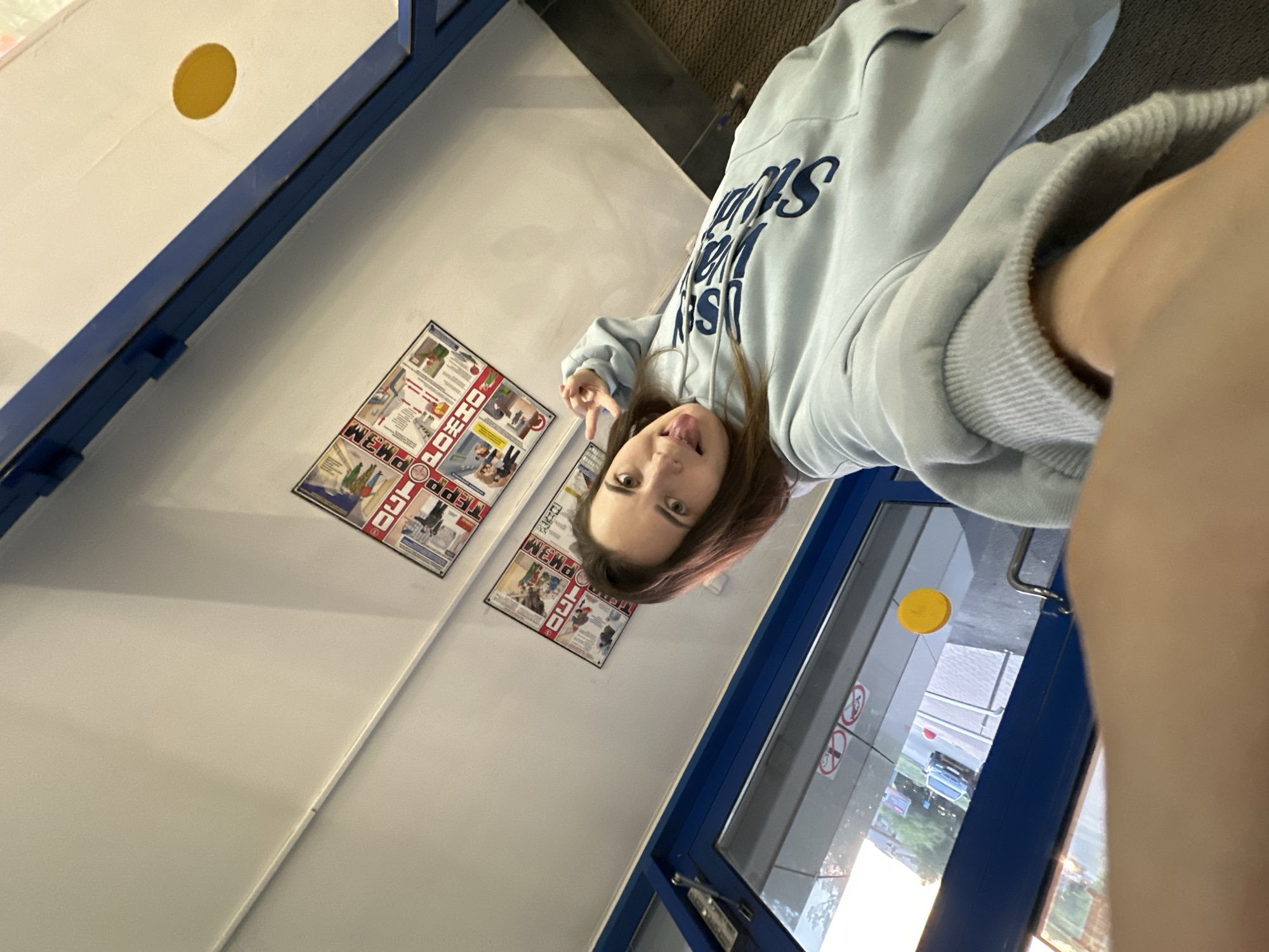 A young person with brown hair and wearing a light blue hooded sweatshirt is taking a selfie while standing inside a building. The person is making a face, sticking their tongue out, and holding up two fingers in a peace sign. A sign in Russian language is posted on a wall. A closed glass door is to the person's right, and a second yellow circle can be seen through the glass door. The location is Perm, Russia.