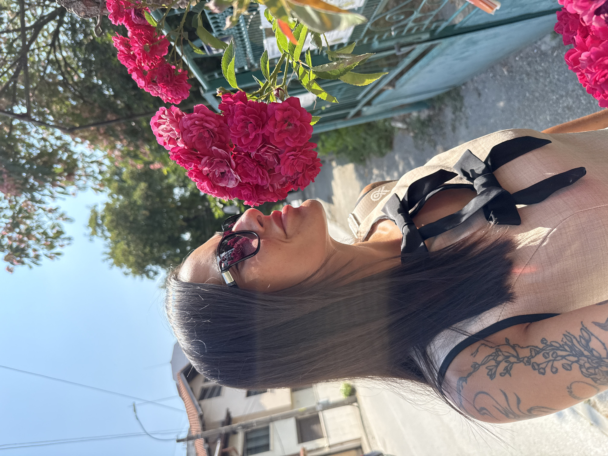 The image shows a woman smelling pink roses outdoors. She has dark hair, sunglasses, and is wearing a light-colored dress with black bows. She has tattoos on her arm. The roses are on a bush next to a green fence. In the background, there are buildings and trees, suggesting an urban setting. The overall scene suggests a sunny day. The image was taken in Pleven, Bulgaria.