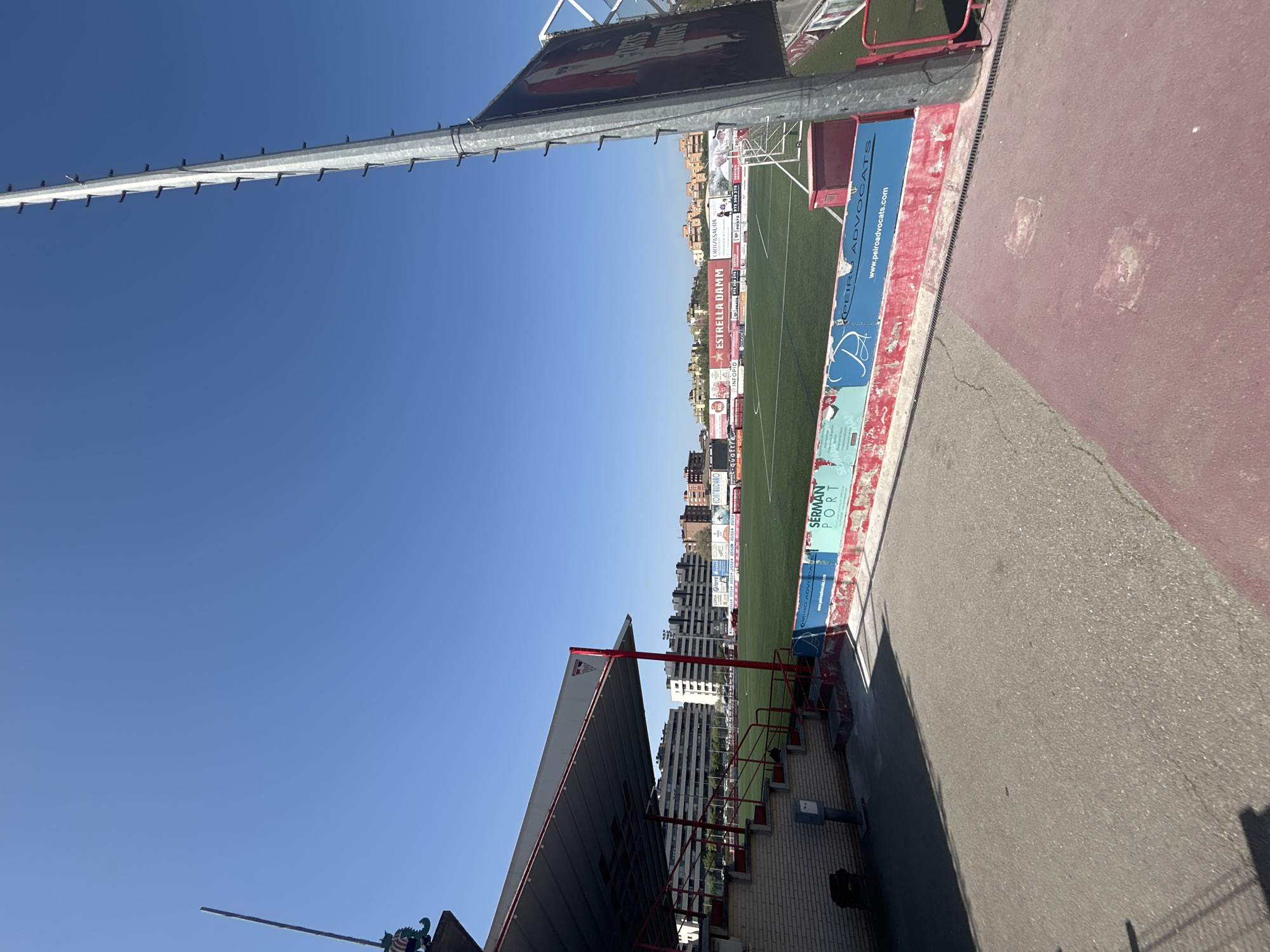 The image depicts a view from a stadium or sports facility in Lleida, Spain. A blue sky is visible above. A tall metal structure extends into the sky. The stadium seating area is to the right, with advertisements for businesses such as Estrella Damm and Serman Port displayed along the wall. Buildings are visible beyond the stadium. A red walkway is also to the right, along with an area of gray concrete. The image is taken at an angle.