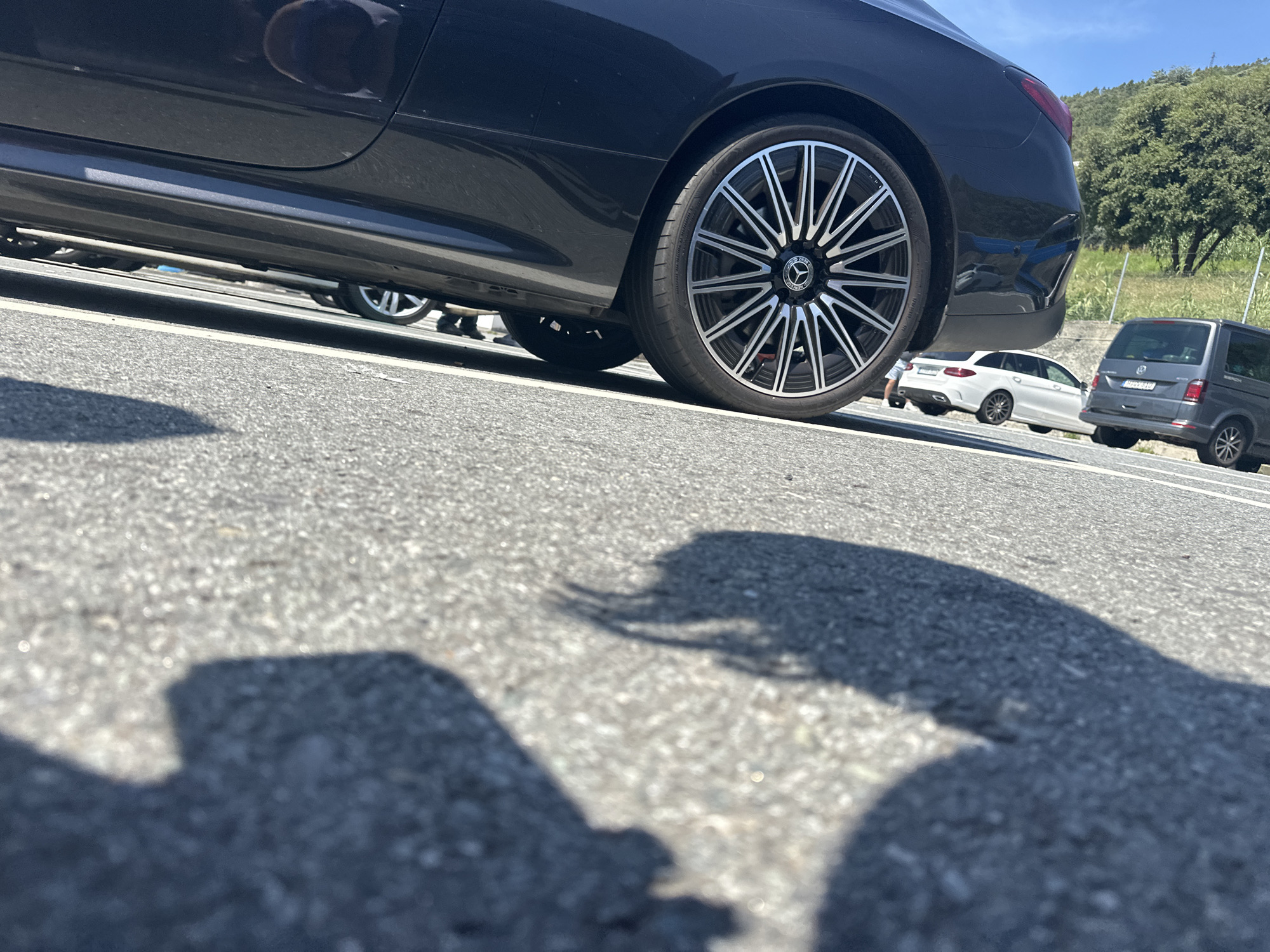 The image shows a parking lot in Varazze, Italy. The angle is low, focusing on the asphalt and the shadows cast by a person taking the picture.
In the background, there are three vehicles:
1. A dark-colored coupe or sedan, with a distinctive multi-spoke wheel design, that seems to be a Mercedes-Benz.
2. A white station wagon of the same brand.
3. A dark gray Volkswagen Transporter van.
The location appears to be a sunny, outdoor parking area. Green vegetation is visible in the distance.