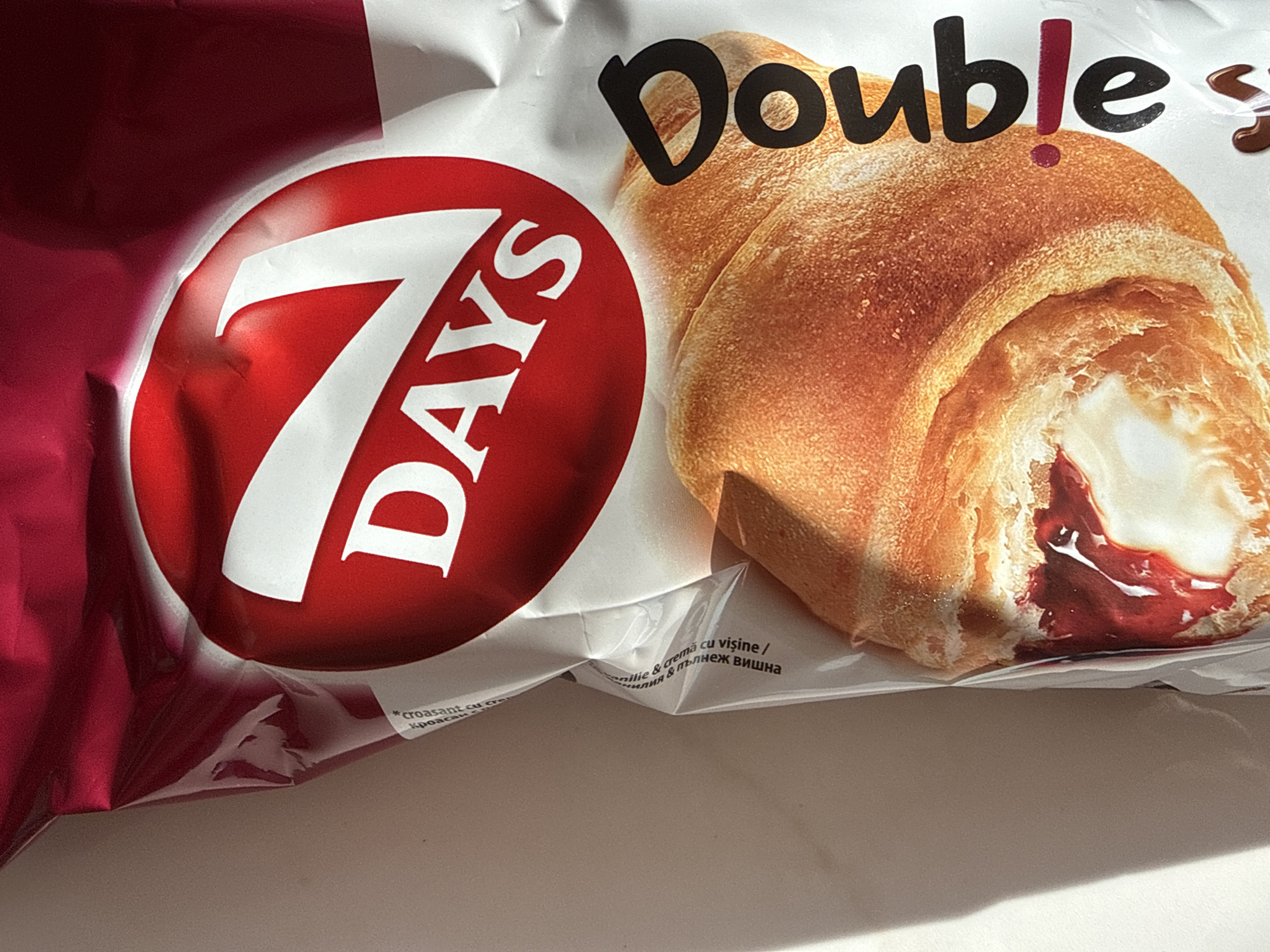 The image depicts a package of "7 DAYS Double" croissants, photographed at a high angle on a plain, light-colored surface in Unknown, Unknown. The packaging features a prominent red oval with "7 DAYS" in white lettering at the top. Below this, the word "Double" is oriented vertically in black with a red dot for the 'i'. An illustration of a golden-brown croissant, cut open to display two distinct fillings—a creamy white substance and a reddish, likely fruit-based filling—occupies the lower portion of the package. Partial text visible on the packaging includes "croasant", "...milie & cremă cu vișine /", and "ния & пълнеж вишна", indicating a cherry or sour cherry cream filling. The package exhibits minor wrinkles. A distinct shadow is cast from the left, suggesting directional lighting. No individuals are present in the scene.