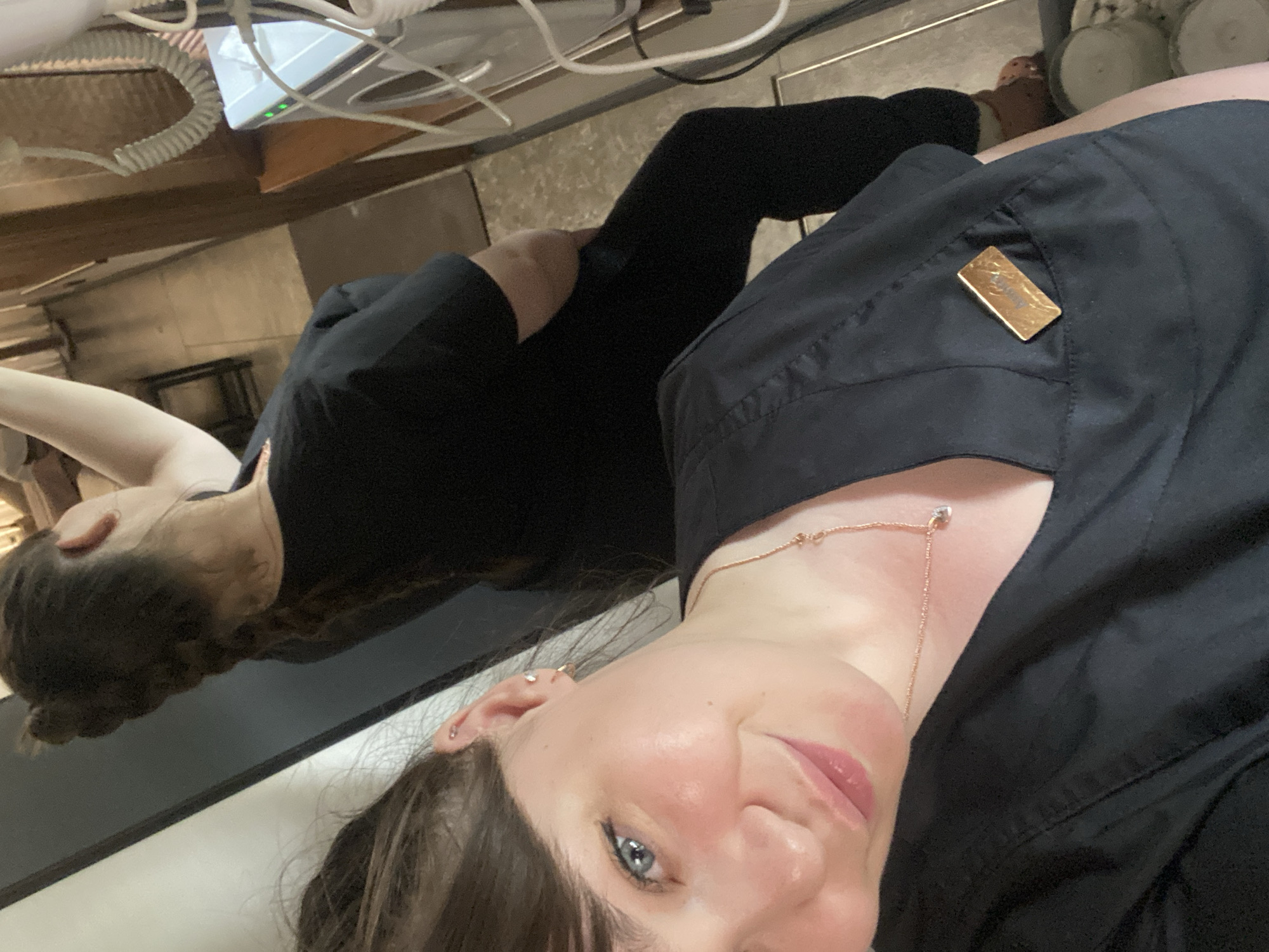 A woman is taking a selfie indoors, wearing a black scrub top. She has brown hair and blue eyes and is wearing a rose-gold necklace and earrings. She appears to be near another person with a similar hairstyle who is wearing a black scrub suit. The background includes medical equipment, possibly in a clinic or dental office. There is no information available to determine if this photo was taken in Votkinsk, Russia.