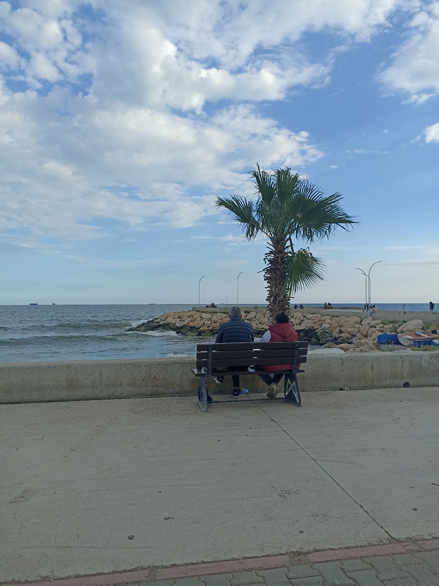 Two people are sitting on a wooden bench facing the sea in Yenişehir, Turkey. A man in a dark jacket and a woman in a red jacket are seated on the bench, positioned in front of a concrete wall. A palm tree is located between the bench and a rocky breakwater with additional people visible on the breakwater. The sea stretches out to the horizon, with ships visible in the distance under a partly cloudy blue sky. The foreground shows a paved surface with a crack running through it and a tiled edge. The lighting suggests it is daytime.