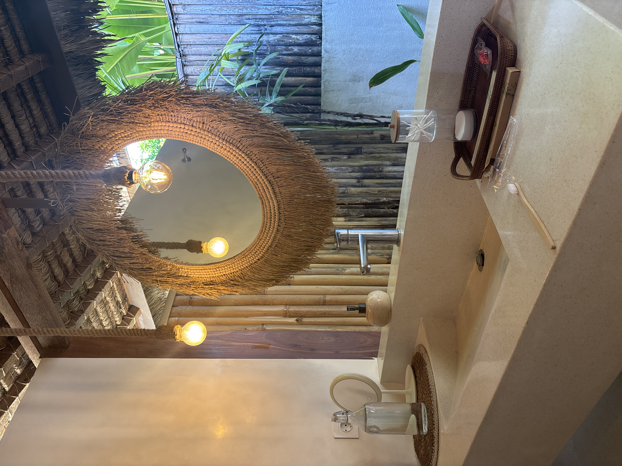 The image captures a bathroom interior in Kuta Selatan, Indonesia. A round mirror with a straw-like fringe hangs above a sink. Three bare light bulbs illuminate the scene, suspended by a thick rope. The sink counter, made of a light-colored material with small speckled details, holds a clear container with cotton swabs, a dark brown woven tray containing toiletries, and a toothbrush. A silver faucet and a soap dispenser are mounted on the countertop. A second round, straw-fringed item rests below the sink, possibly a mat. Above the mirror, glimpses of outdoor greenery and a textured bamboo-like wall can be seen. The roof is constructed from straw or thatch.
